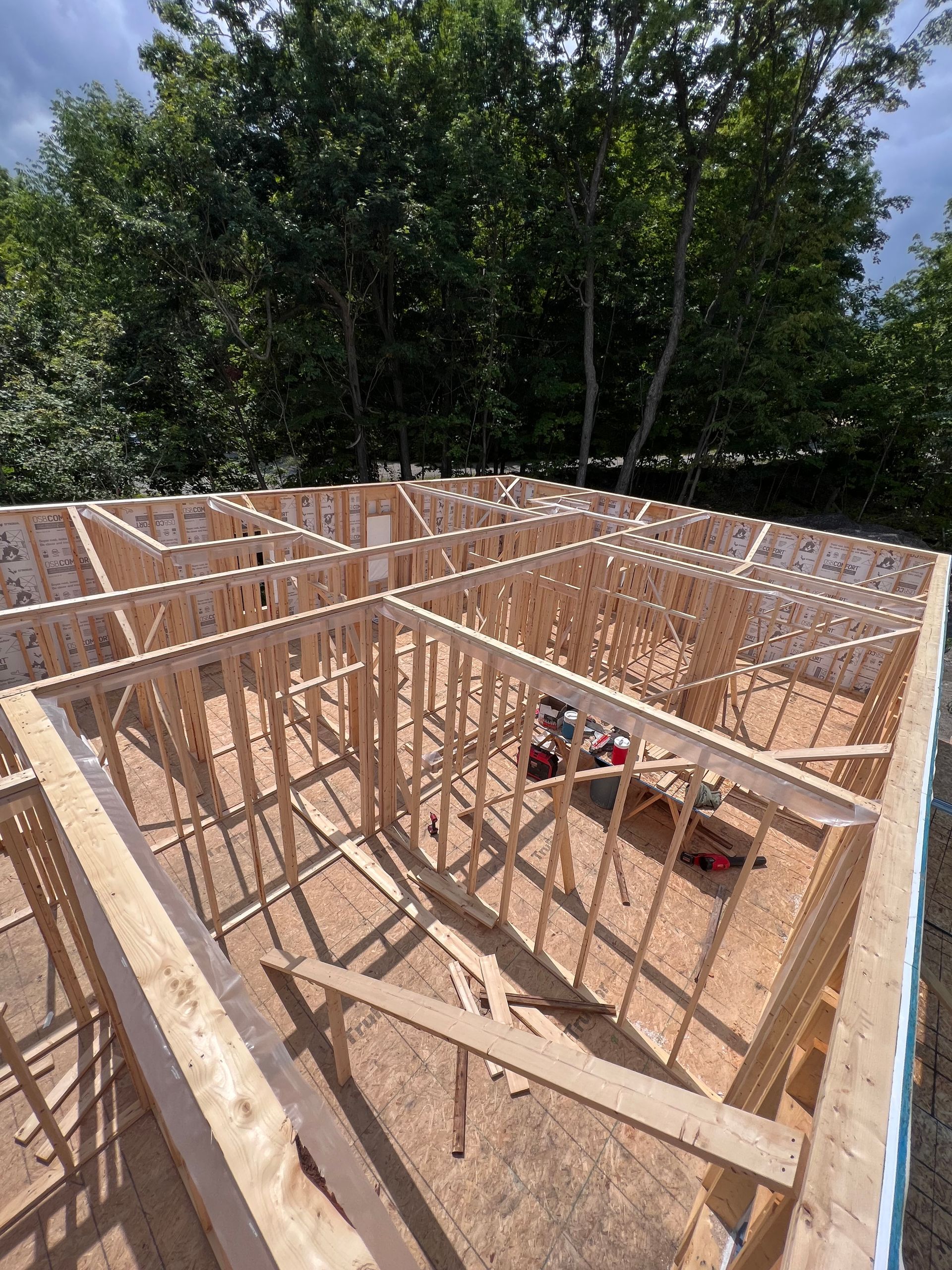 Maison à ossature bois en construction, avec montants apparents et plafond ouvert, entourée d'arbres.