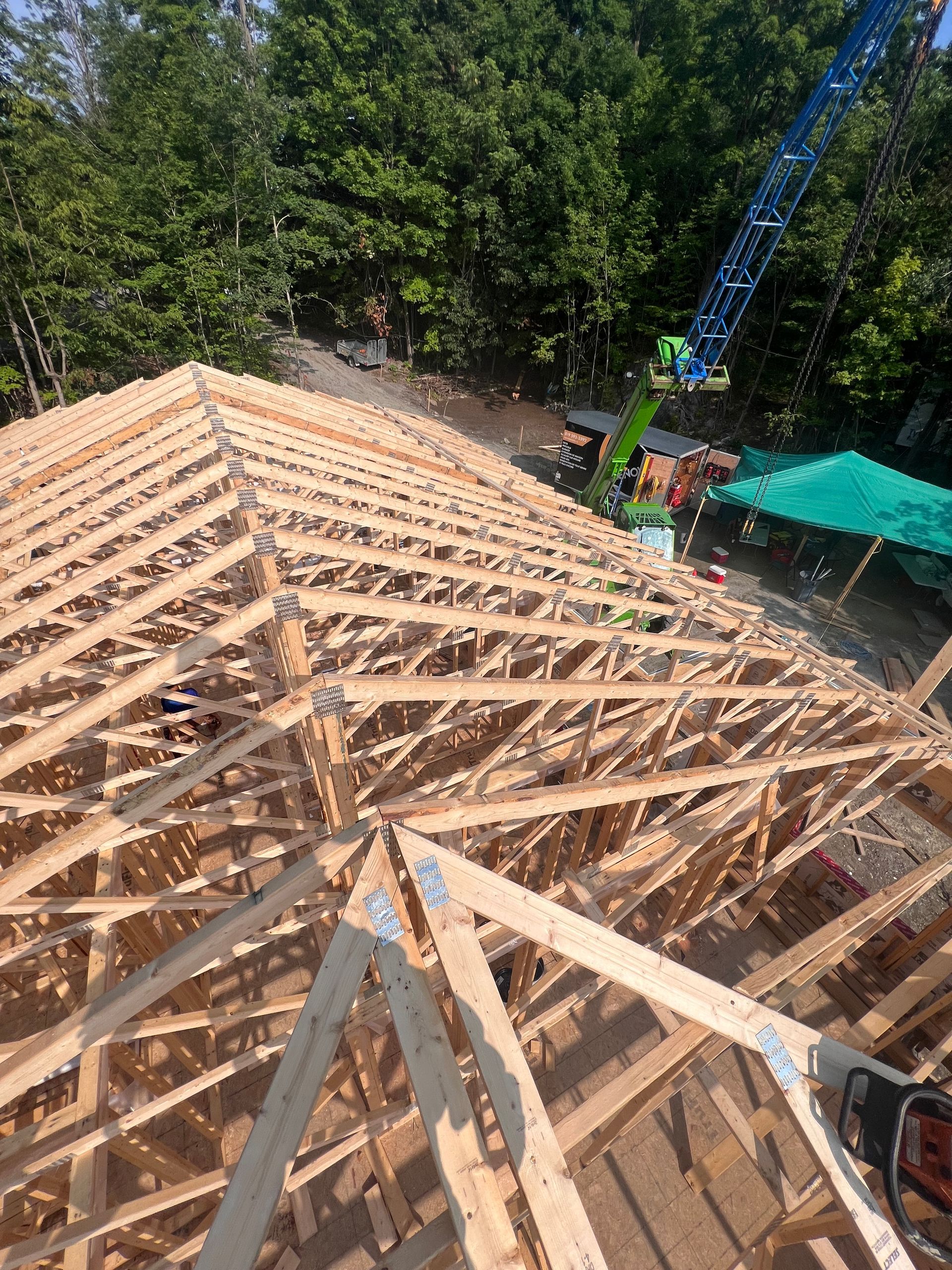 Charpente de toit en construction sur une maison, avec une grue, une tente verte et des arbres en arrière-plan.
