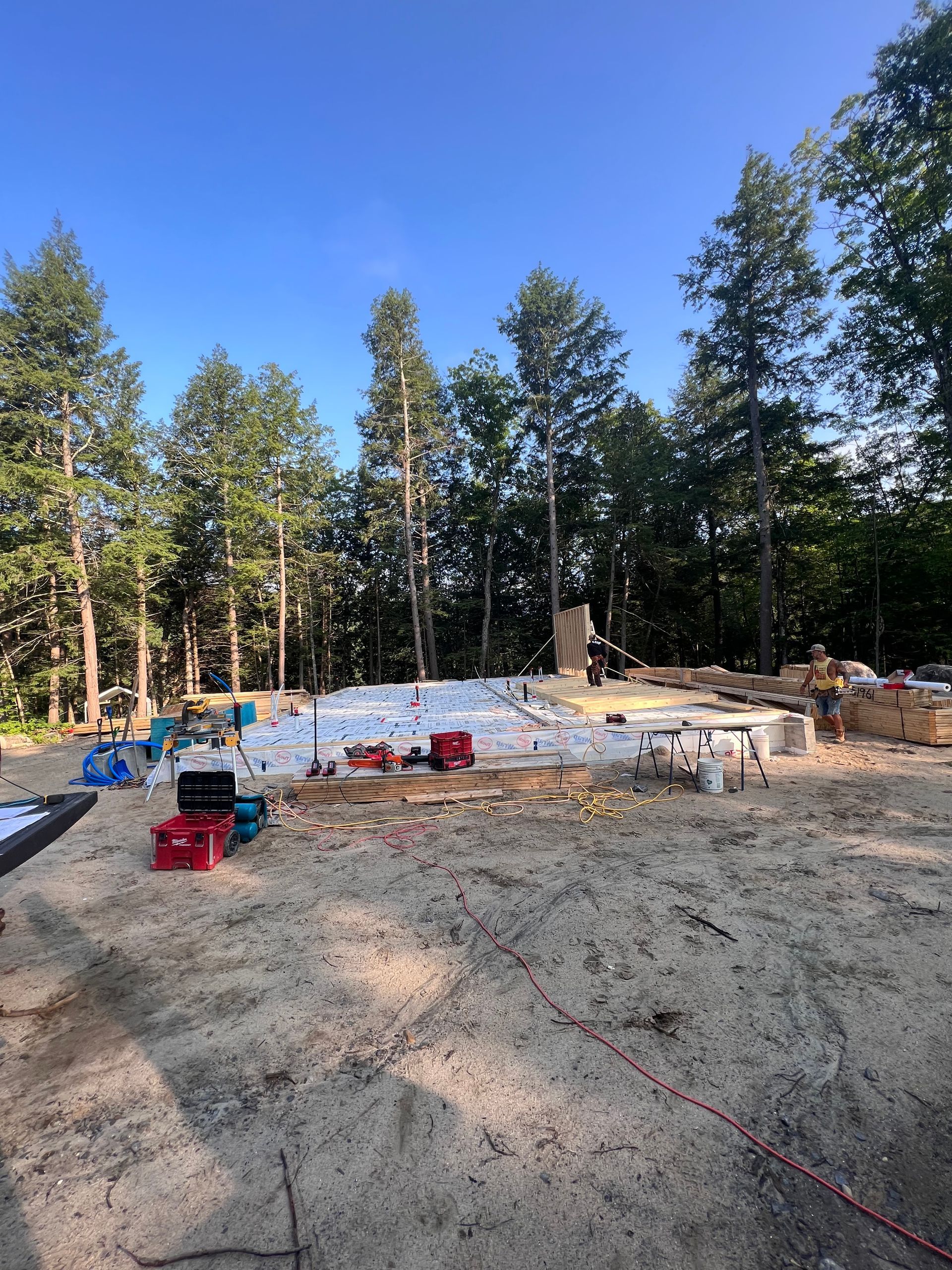 Chantier de construction en plein air avec des matériaux de construction, des caisses rouges et une bâche blanche dans une clairière entourée de grands arbres