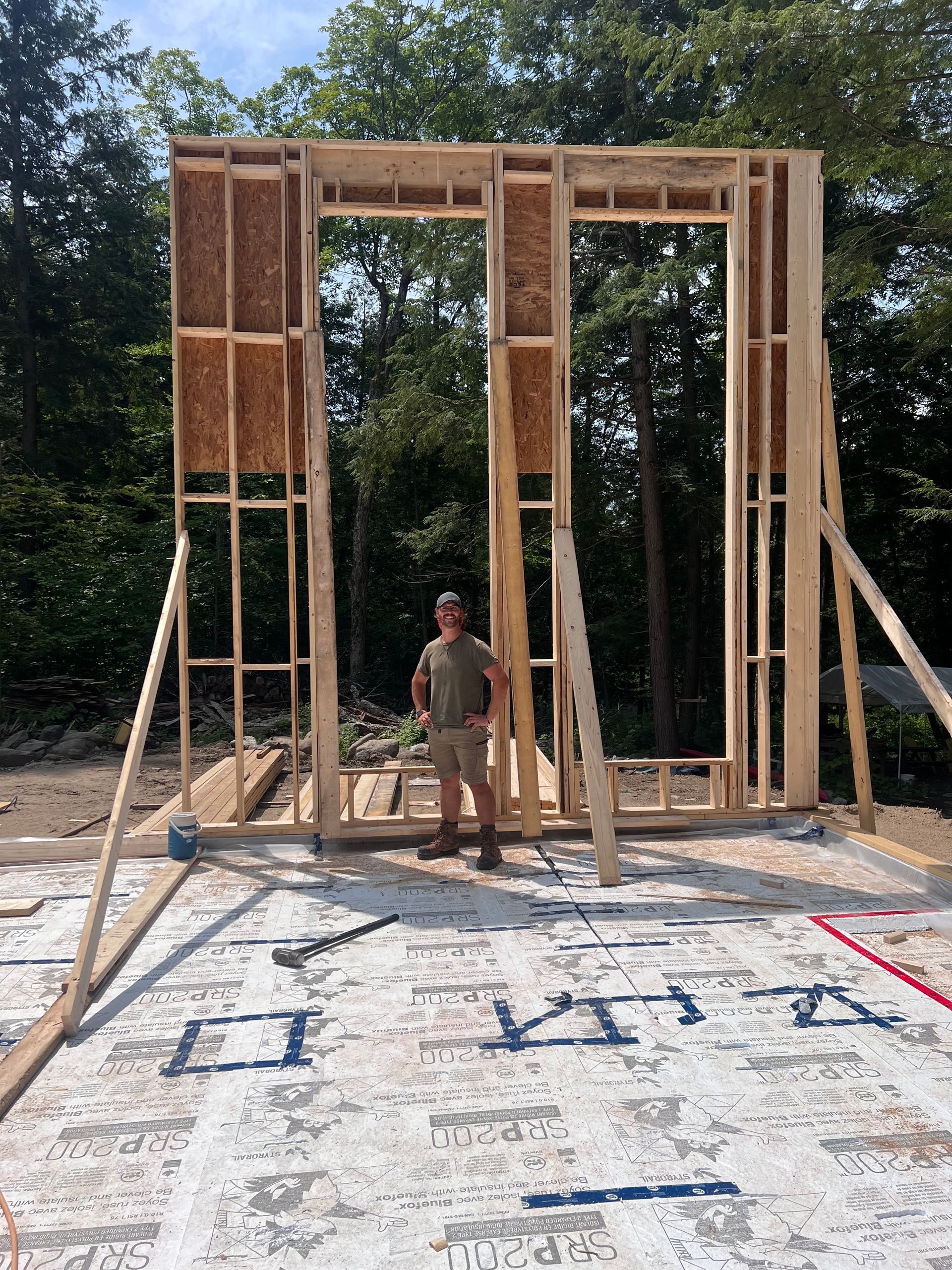 Charpente en bois d'une maison en construction, avec une personne debout dans l'embrasure de la porte, sur une dalle de fondation.