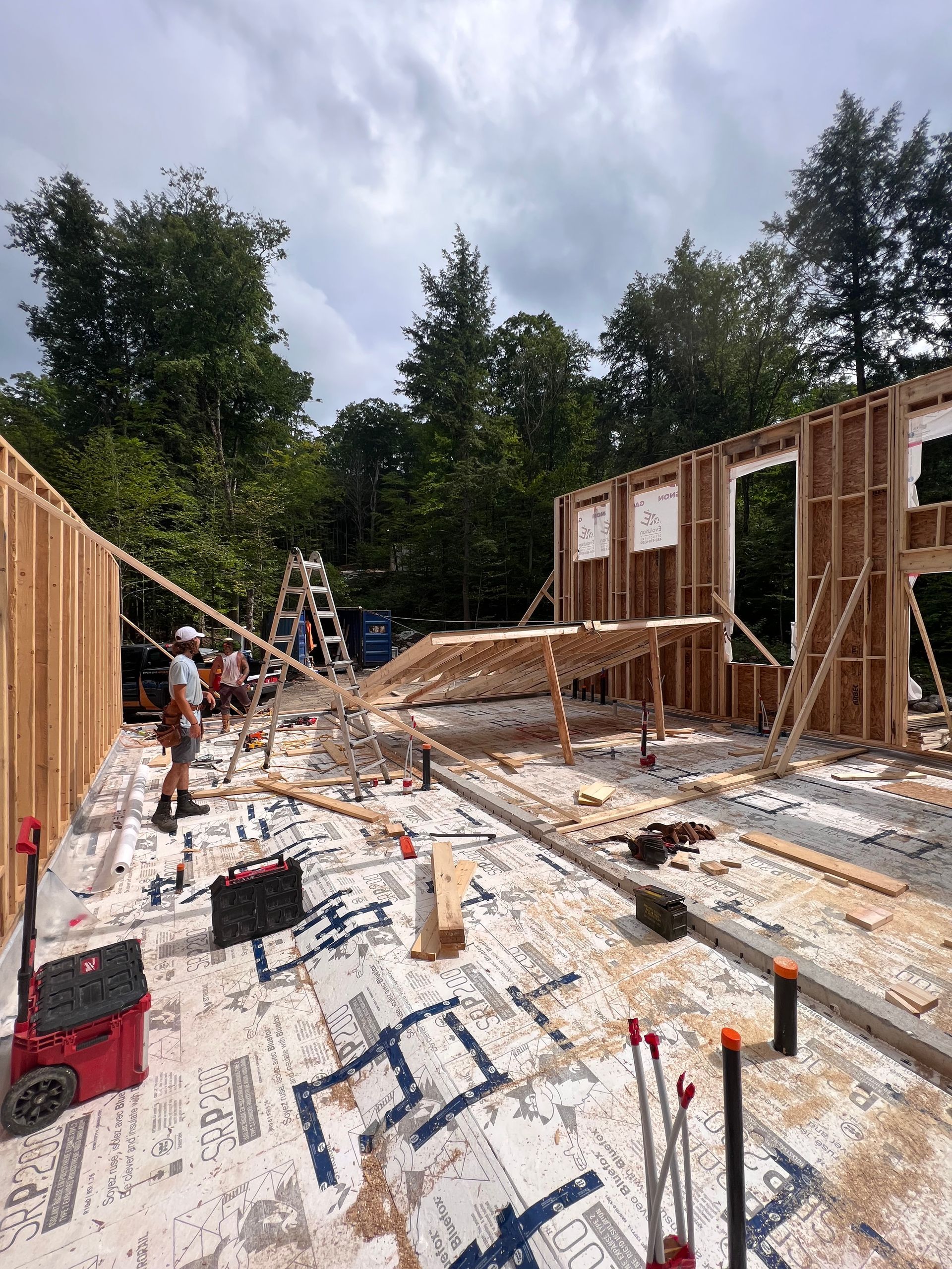 Maison à ossature bois en construction, avec des ouvriers, des outils et du bois sur un plancher en contreplaqué sous un ciel nuageux.