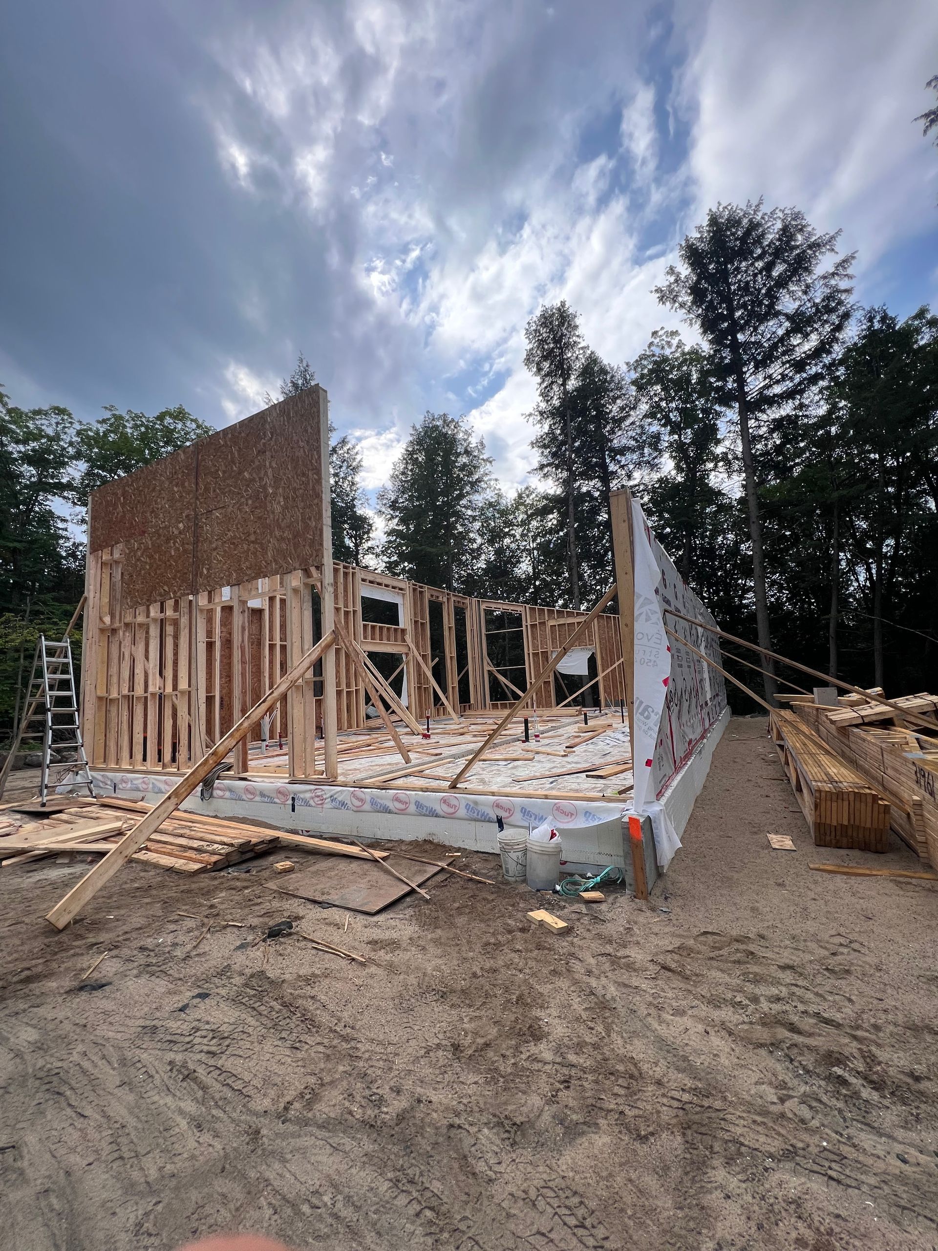 Maison partiellement en construction sur un terrain vague, avec des murs en bois apparents et un ciel nuageux.