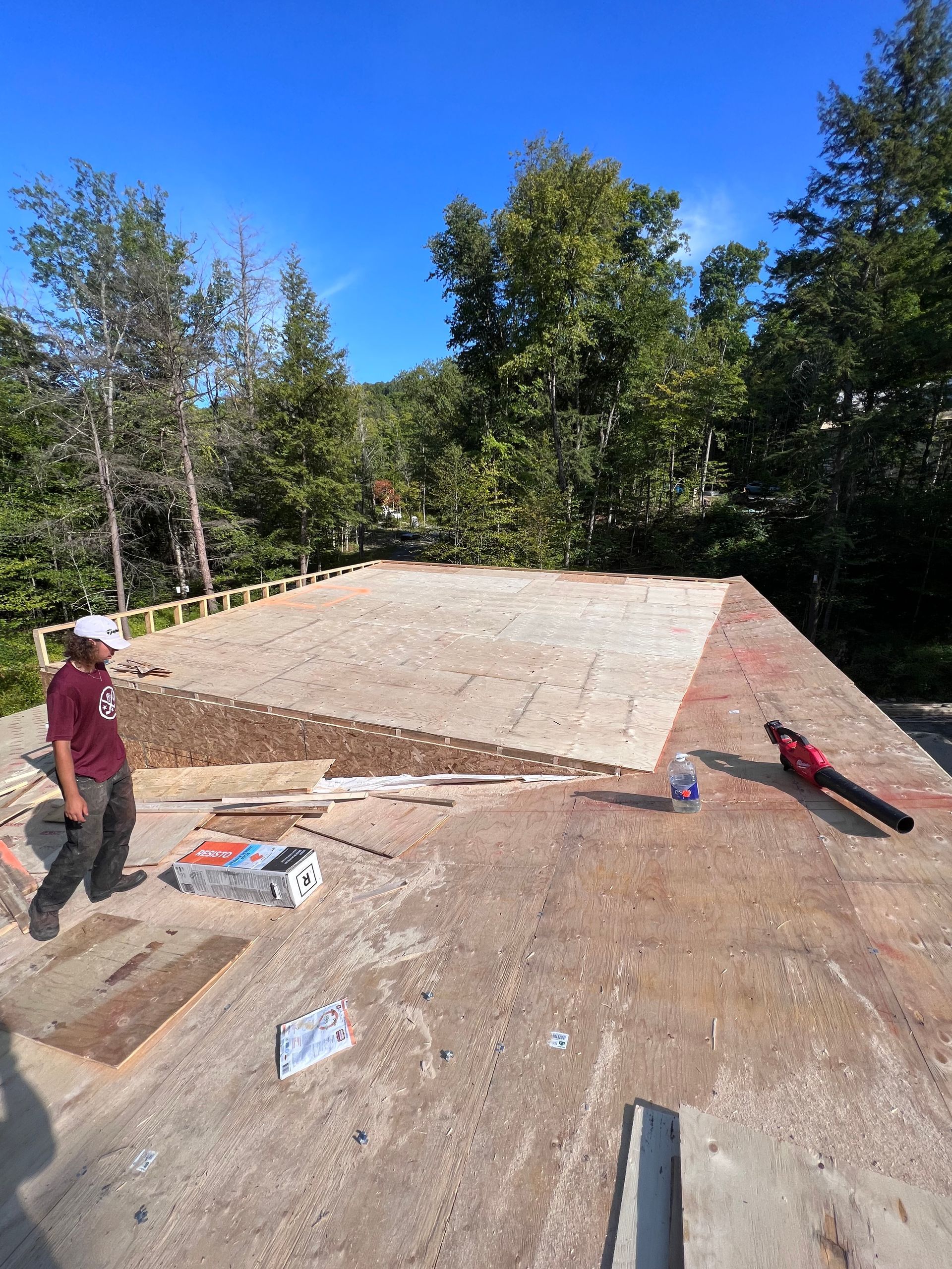 Des ouvriers posent des bardeaux sur le toit d'une petite structure située sur un site boisé et ensoleillé.