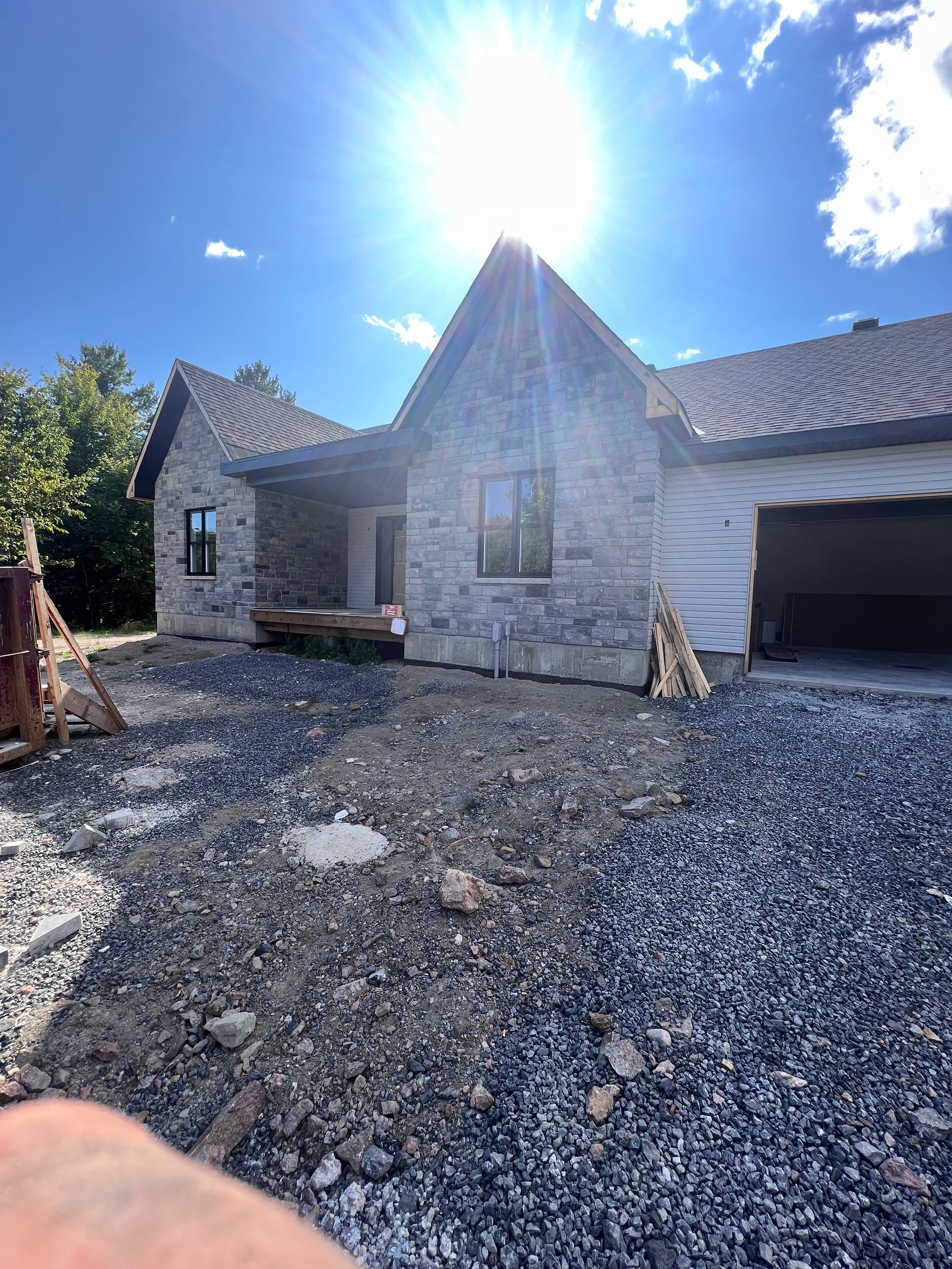Maison en pierre en construction avec cour gravillonnée, soleil éclatant et garage attenant.