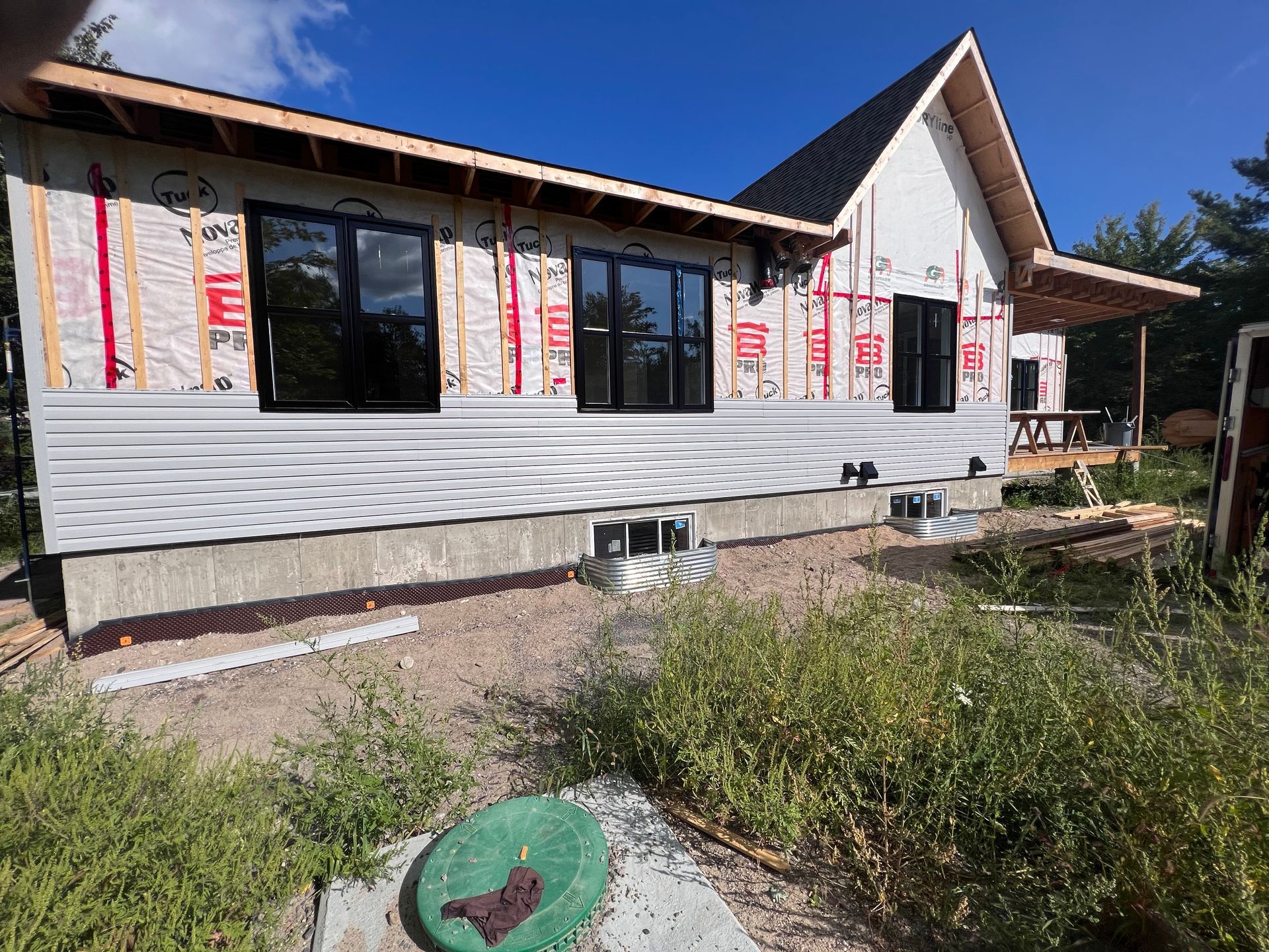 Maison partiellement construite, avec un bardage blanc et des fenêtres noires, entourée de mauvaises herbes sous un ciel bleu clair.