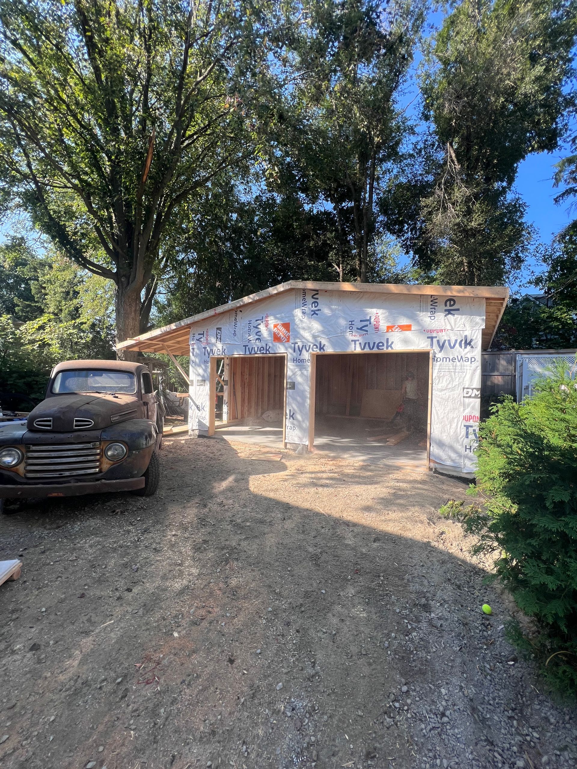 Garage rustique avec deux travées ouvertes et un camion garé sous de grands arbres, au bout d'une allée de terre.