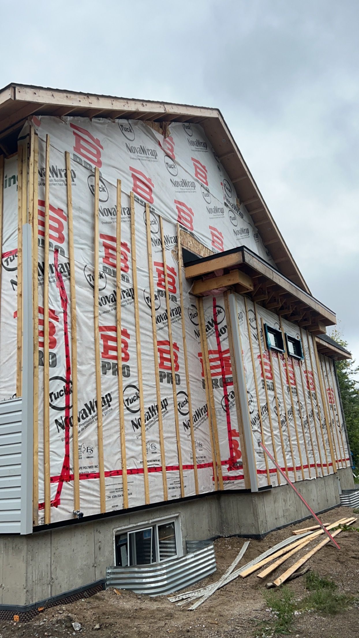 Maison en construction avec murs extérieurs recouverts d'un revêtement, charpente apparente et bois empilé à côté.