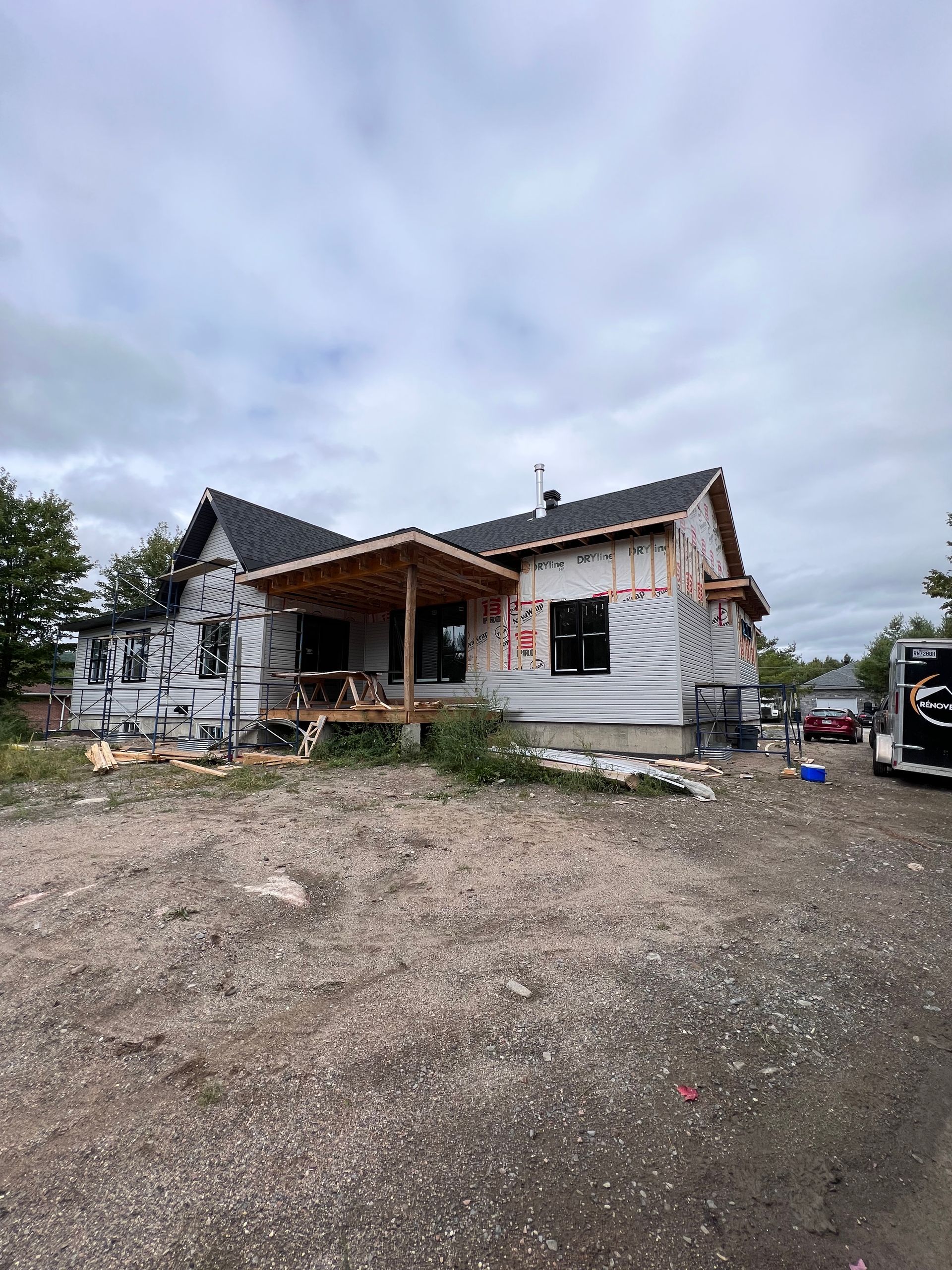 Maison blanche partiellement démolie, charpente apparente et débris sur un terrain de gravier sous un ciel nuageux