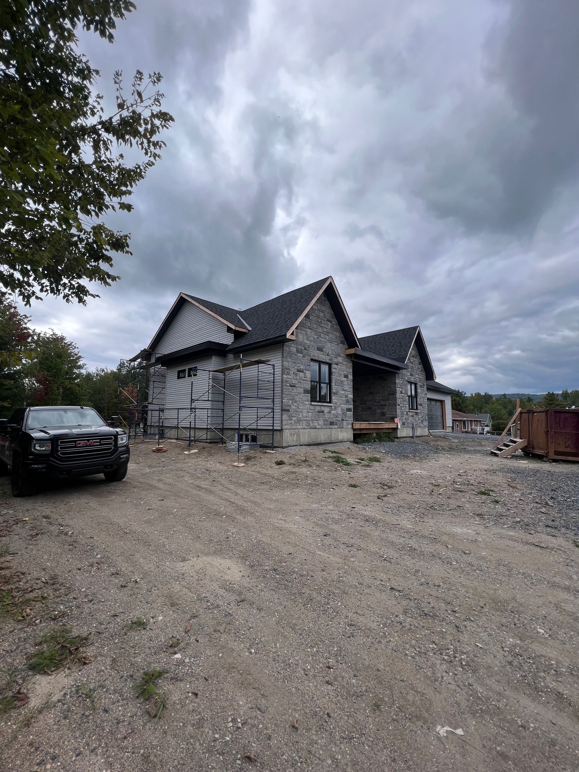 Chantier de construction nuageux avec une maison en construction, un terrain gravillonné et une voiture noire garée