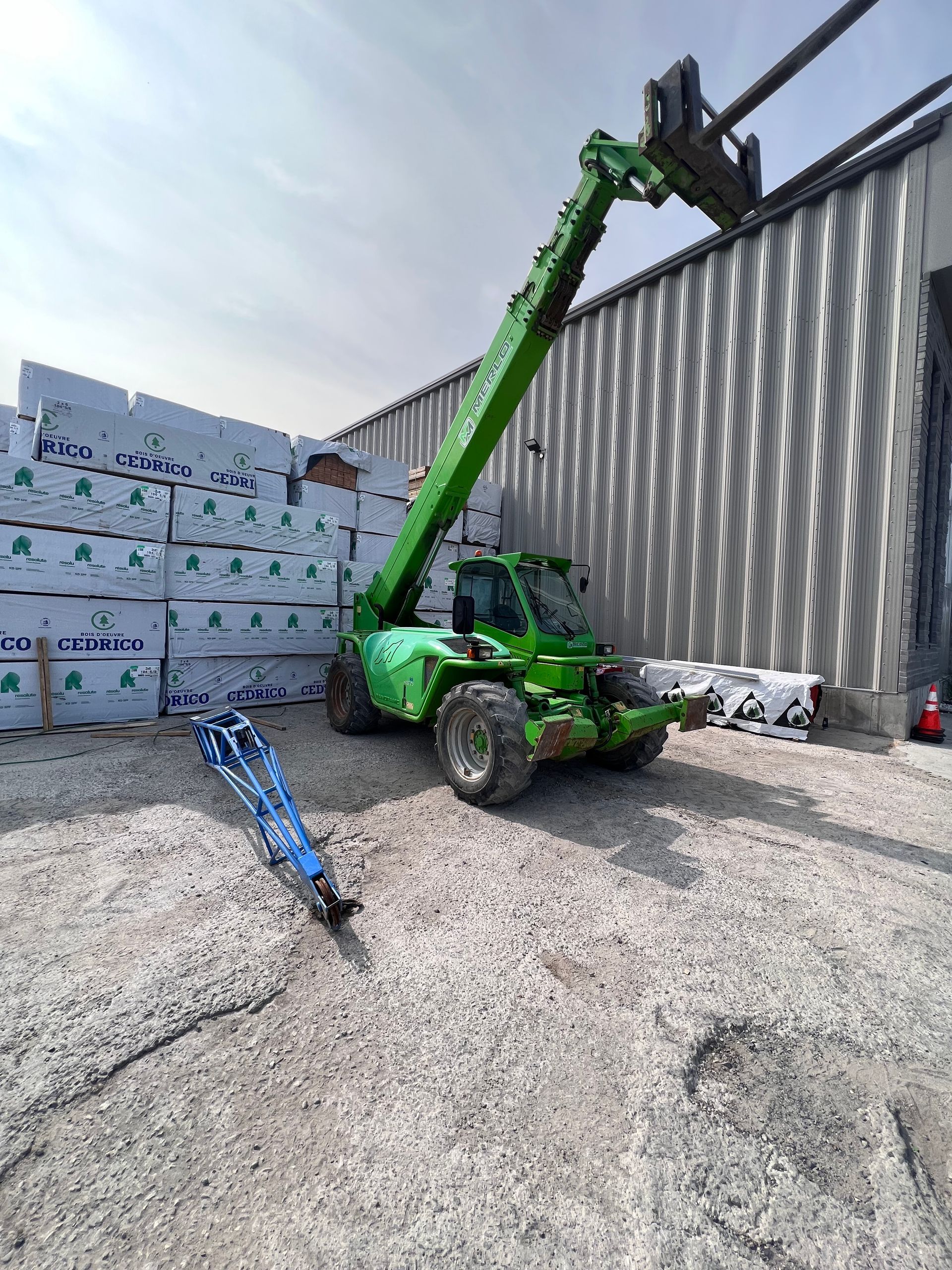 Un chariot télescopique vert soulève des matériaux à côté de palettes empilées et d'un mur d'entrepôt en carton ondulé.
