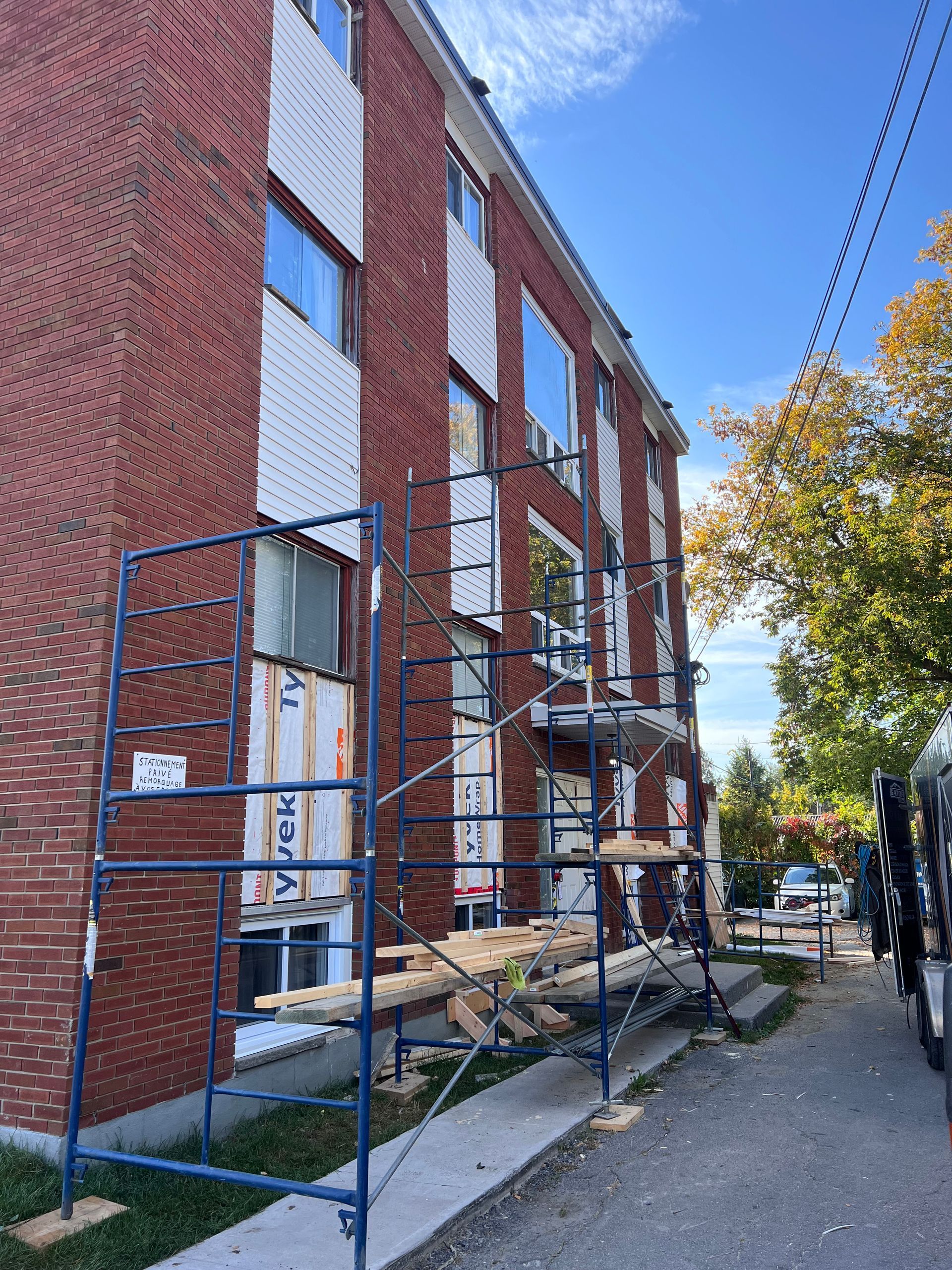 Échafaudages à côté d'un bâtiment en briques rouges sur un trottoir, avec des arbres et des voitures garées en arrière-plan