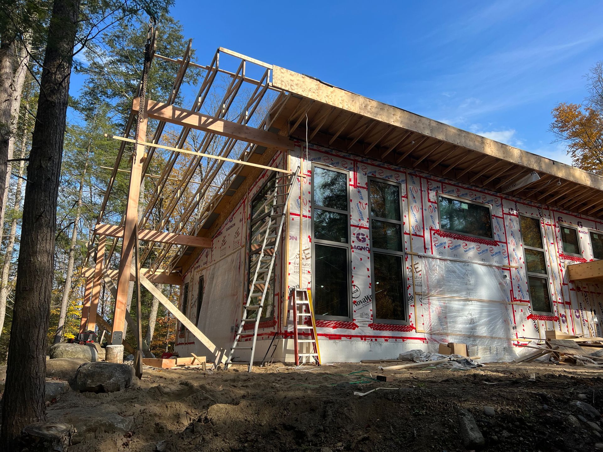 Maison en construction, partiellement bâtie, avec des échafaudages et de grands pins à proximité.