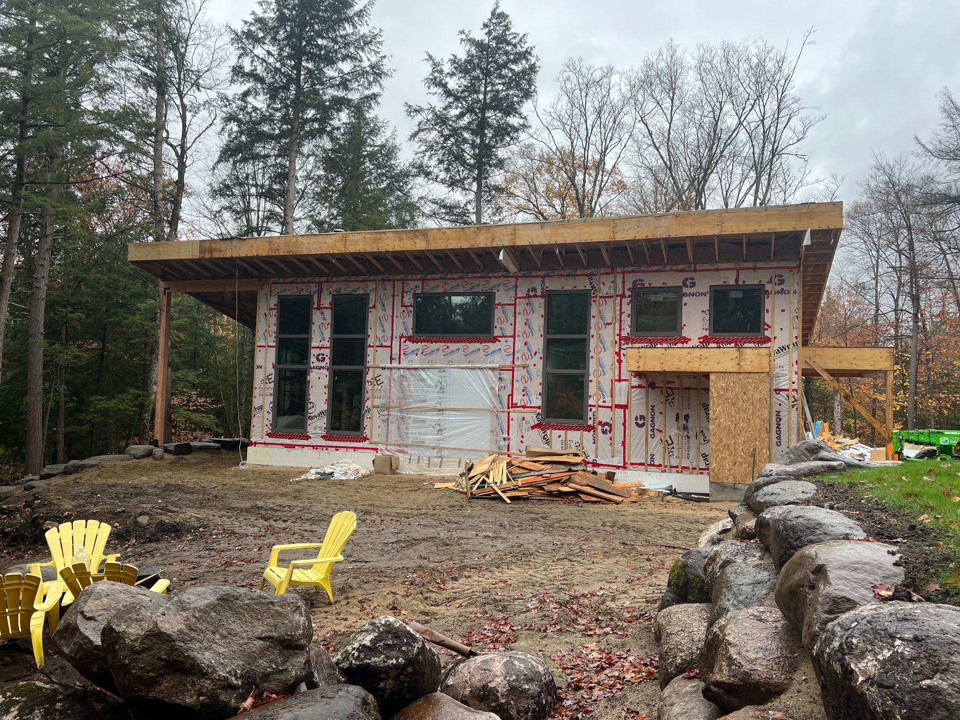 Maison partiellement construite avec isolation apparente, entourée d'arbres, de rochers et de chaises jaunes dans une cour