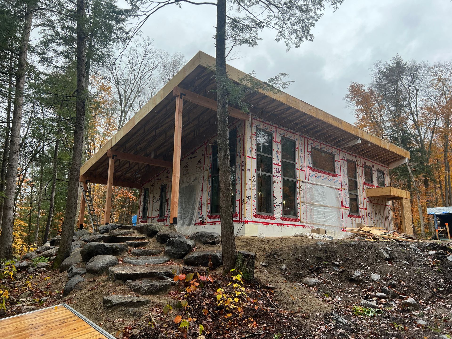 Cabane rustique en construction dans un cadre boisé automnal, avec des marches en pierre et des fenêtres aux encadrements rouges