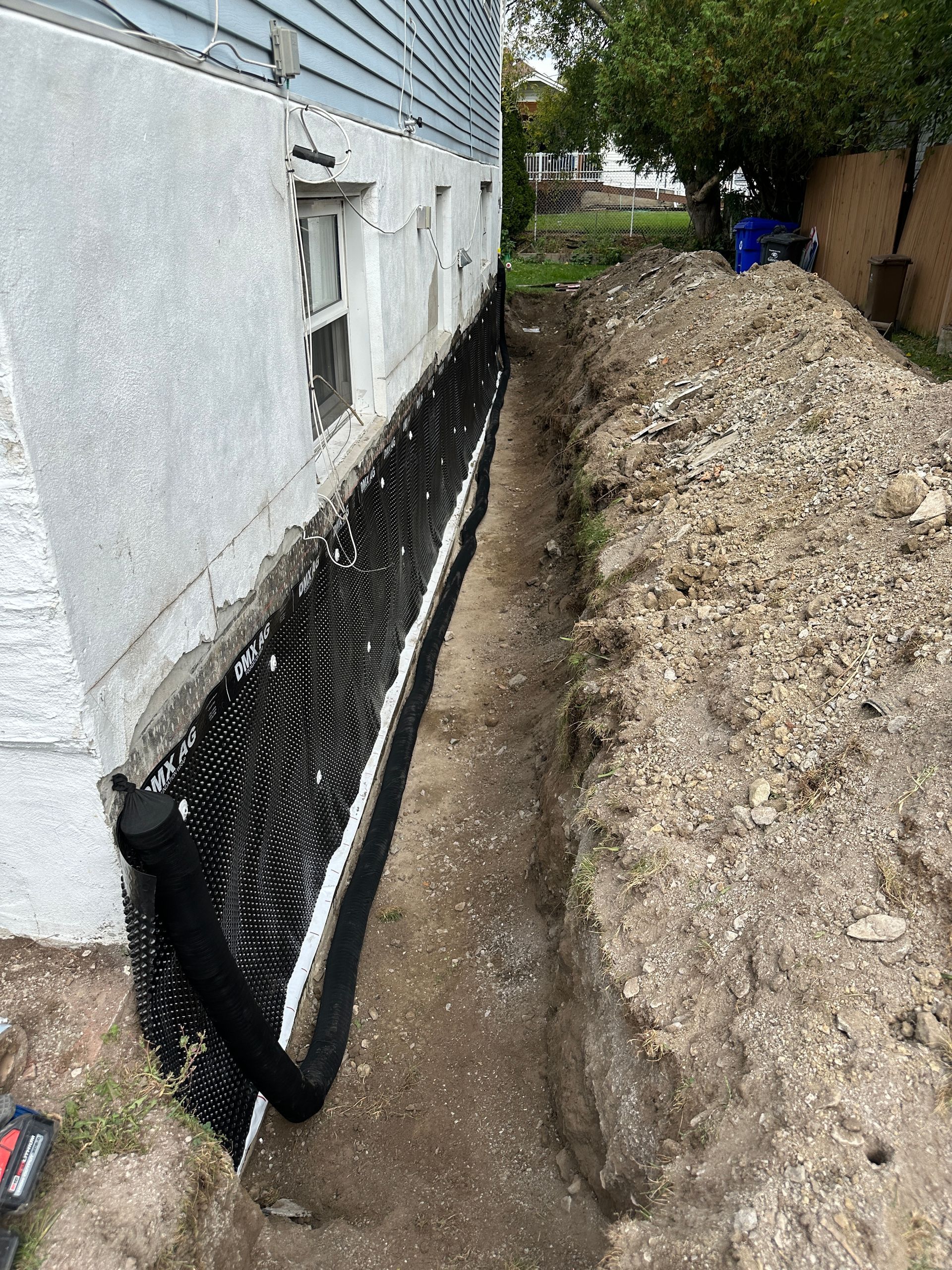 Zone de réparation des fondations à côté d'une maison, avec treillis de drainage noir et tranchée le long du mur.