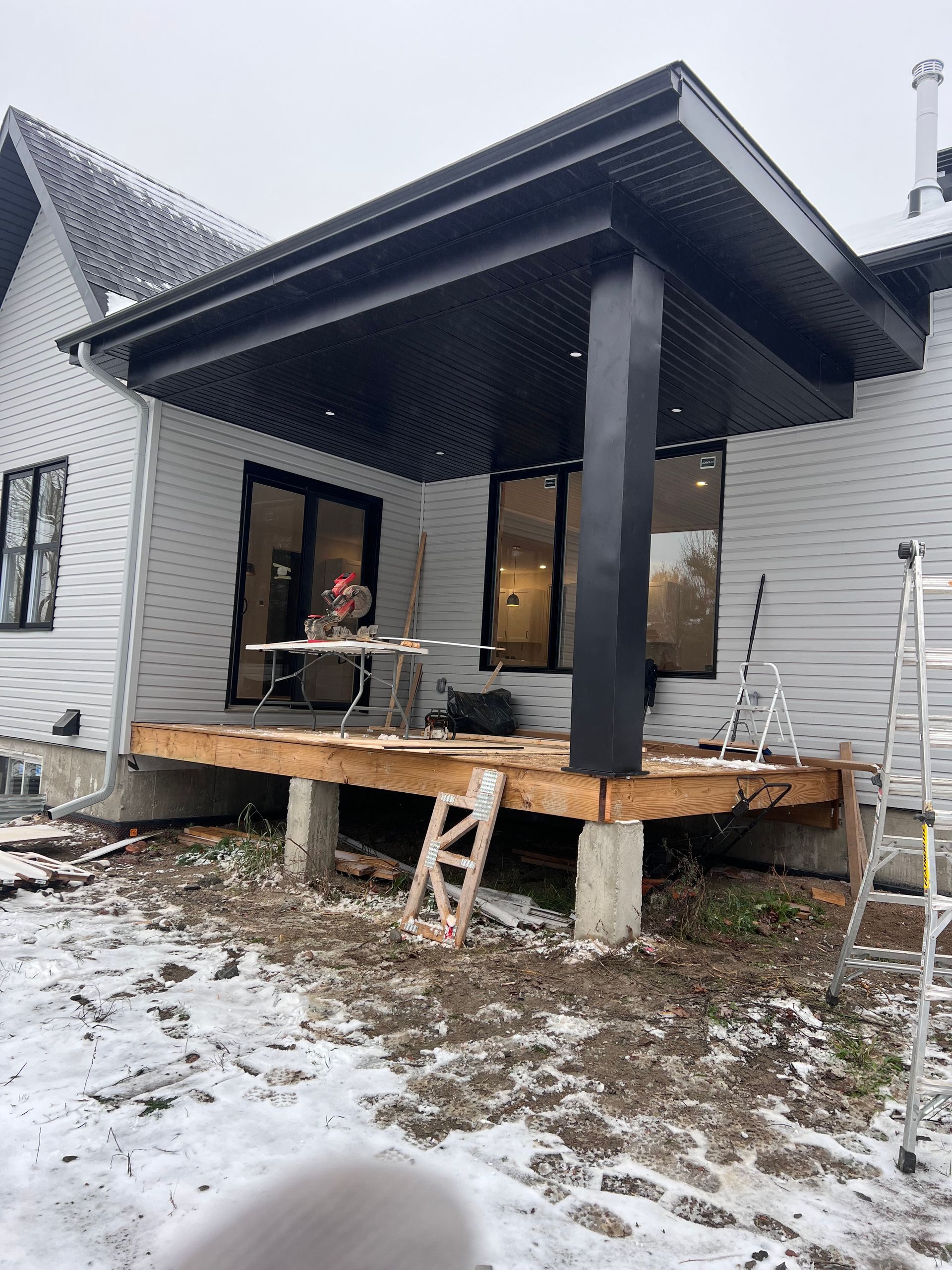 Terrasse enneigée en construction dans le jardin, avec des poteaux noirs et une petite échelle en dessous
