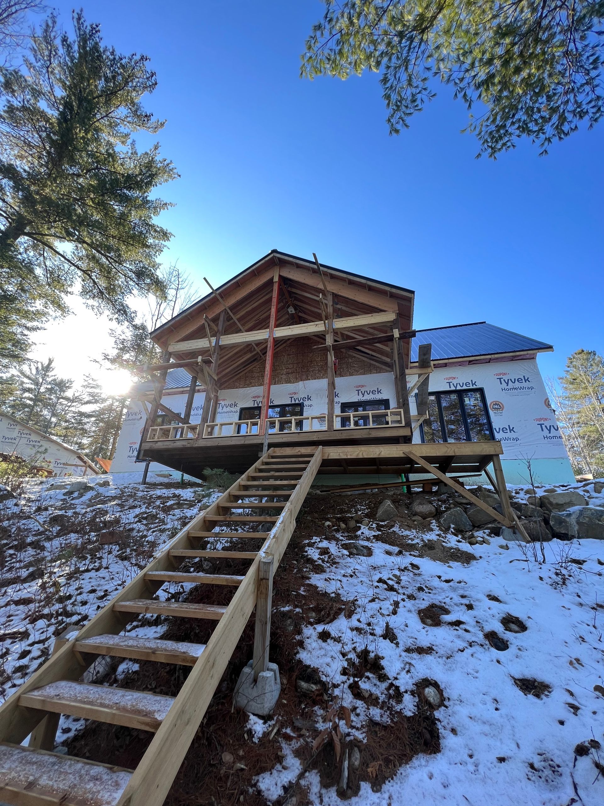 Chalet enneigé à flanc de colline avec un escalier en bois menant à une véranda couverte sous un ciel bleu azur.
