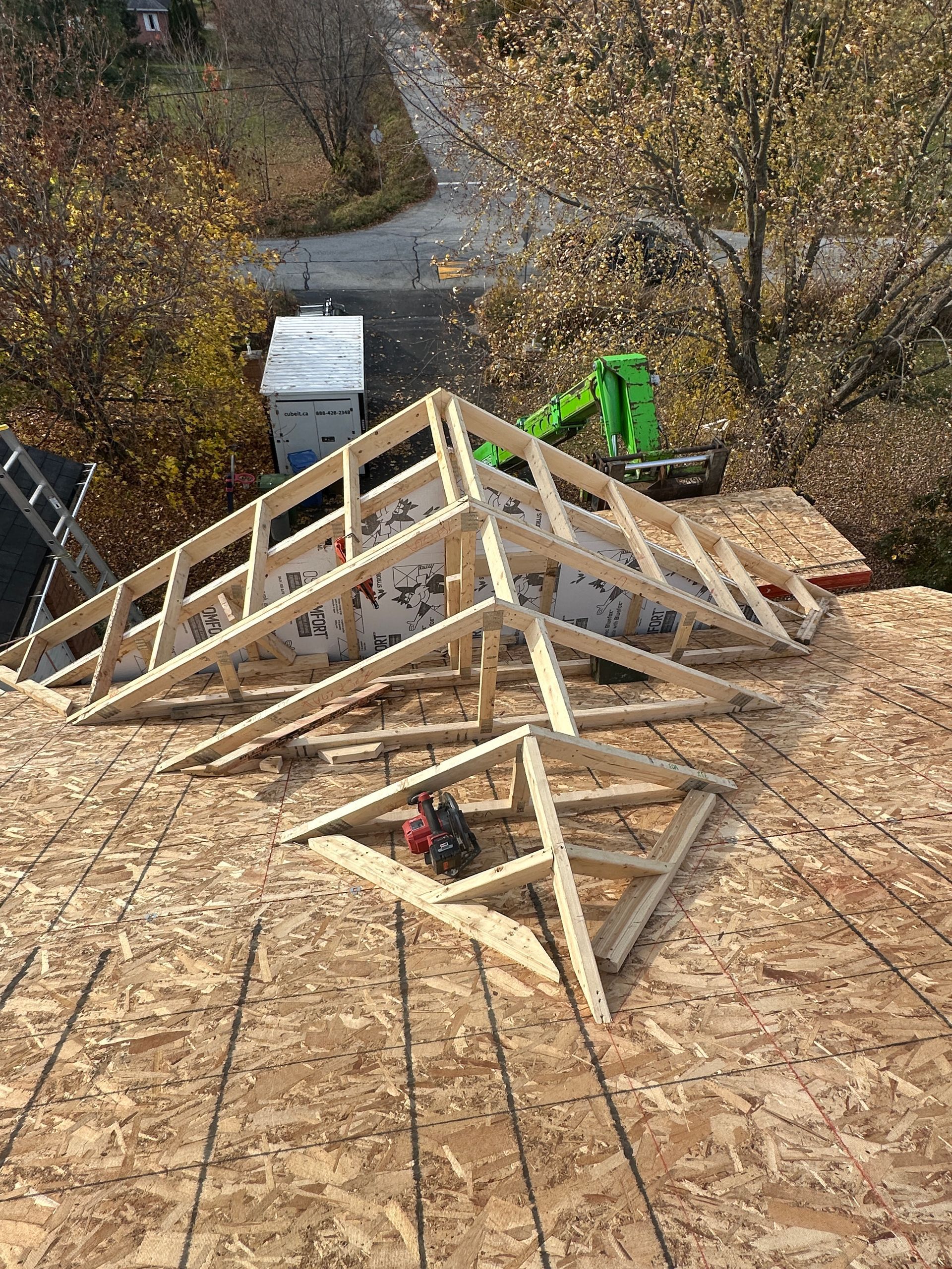 Charpente de toit en bois partiellement construite sur une maison en construction, avec une excavatrice verte à proximité.