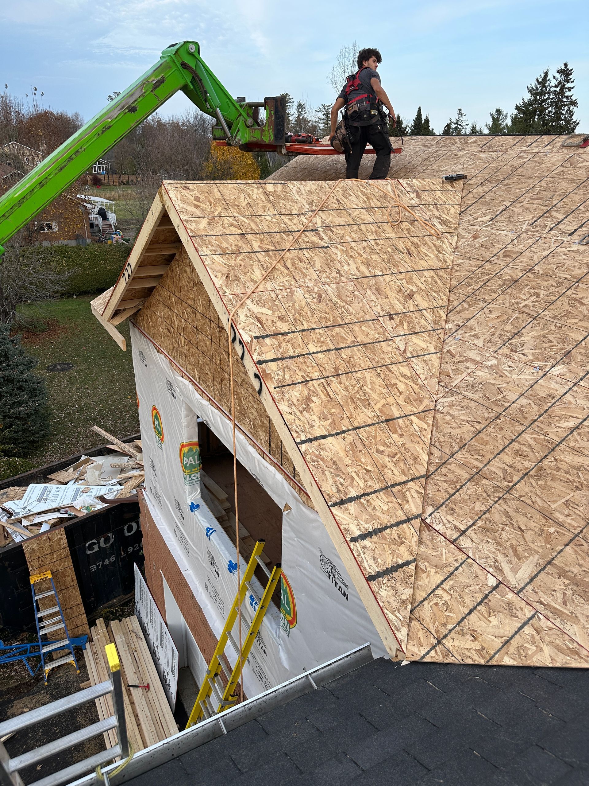 Ouvrier sur un toit en construction, installant un revêtement en contreplaqué à côté du bras vert d'une excavatrice.