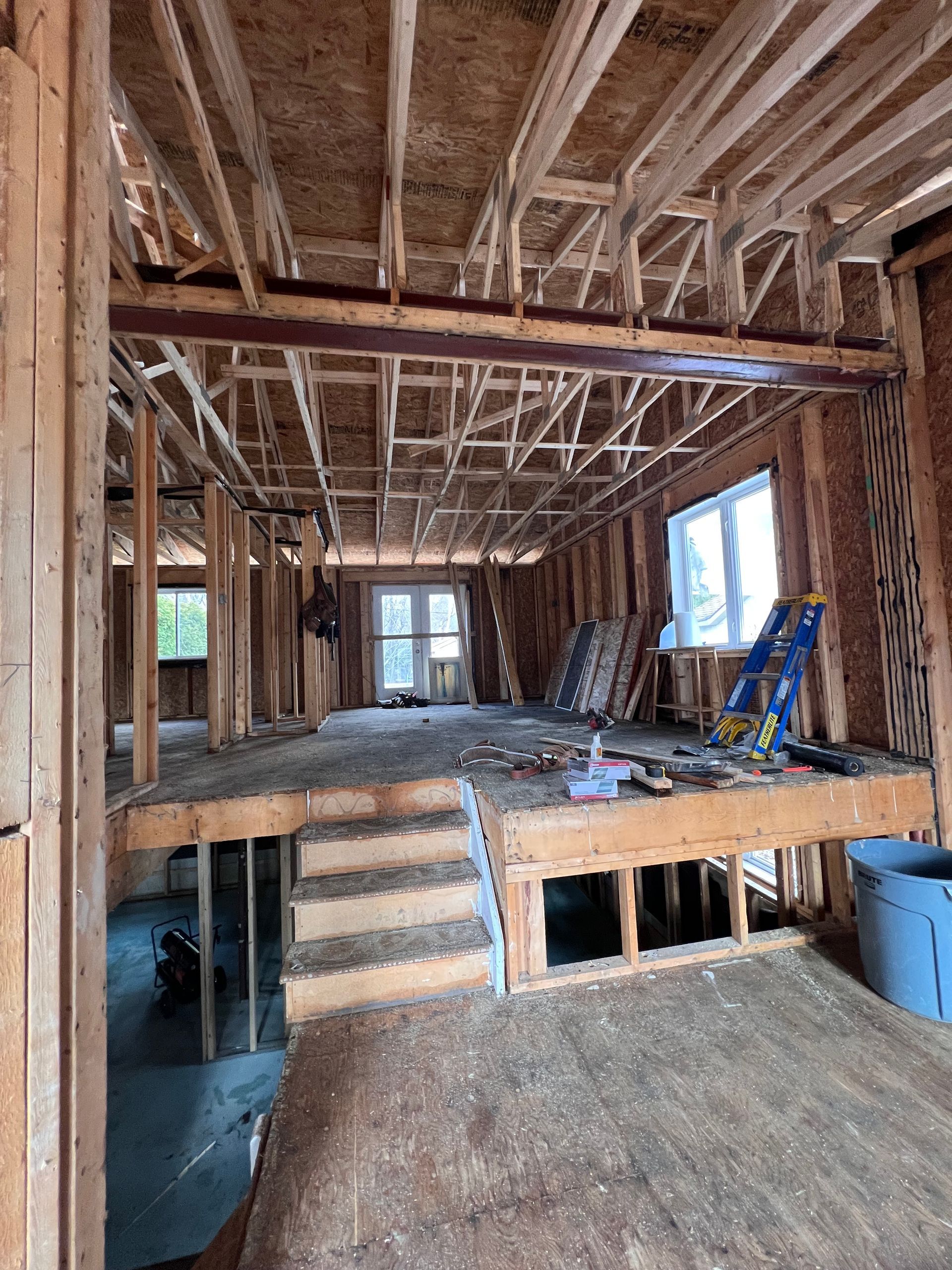 Intérieur d'une maison en construction avec charpente apparente, escalier et échelle bleue près de grandes fenêtres.