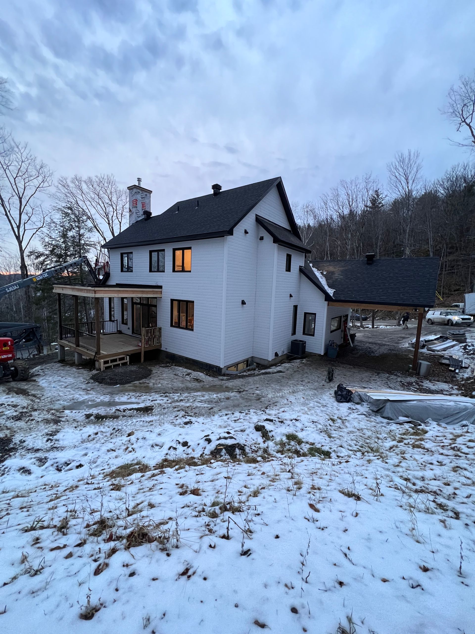 Maison blanche saupoudrée de neige, aux fenêtres éclairées au crépuscule, entourée d'arbres dénudés et d'une cour boisée