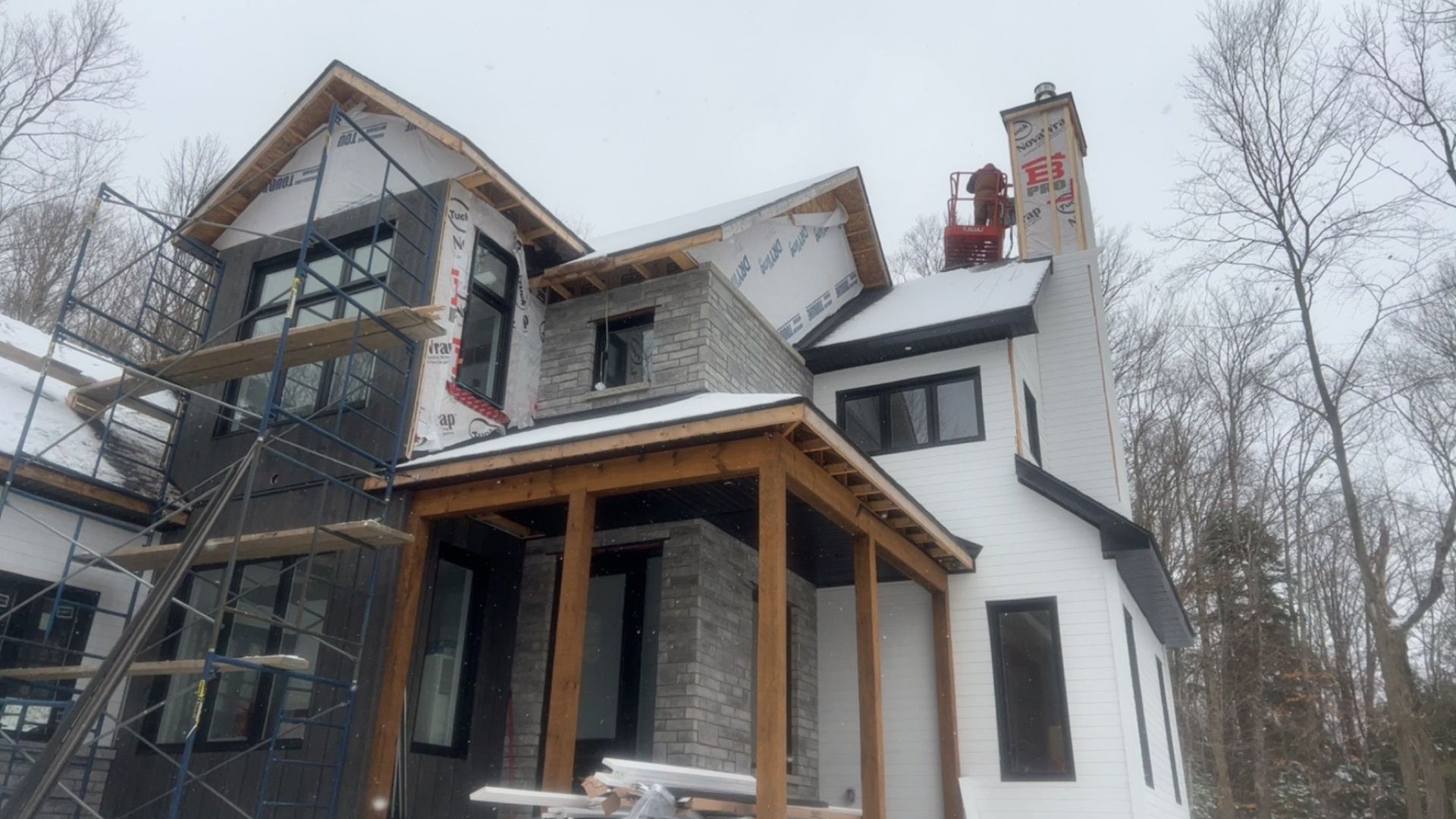 Maison blanche inachevée recouverte de neige, avec des boiseries noires et un porche couvert, dans une zone boisée.