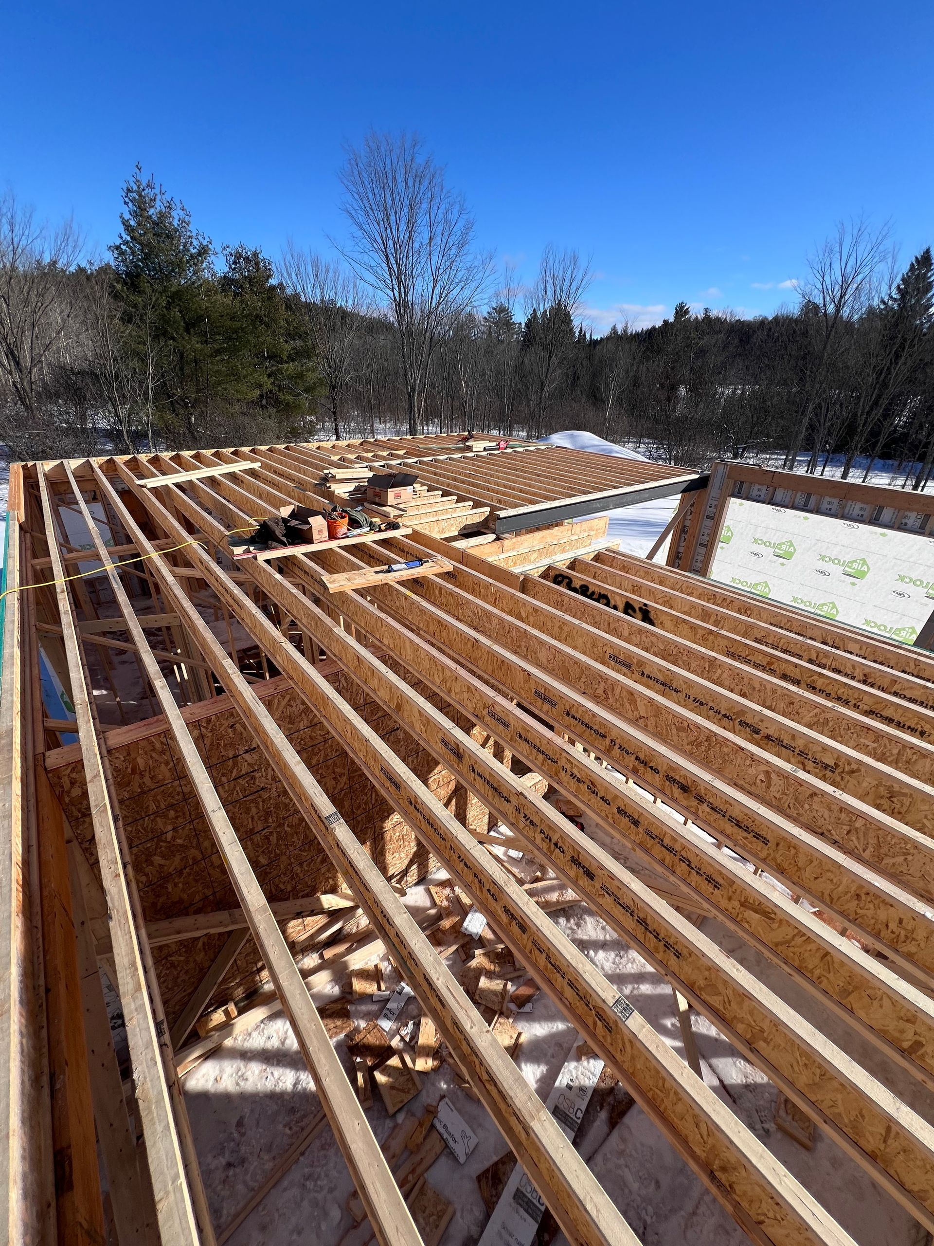 Charpente apparente d'un toit en construction, avec ciel bleu et arbres en arrière-plan.