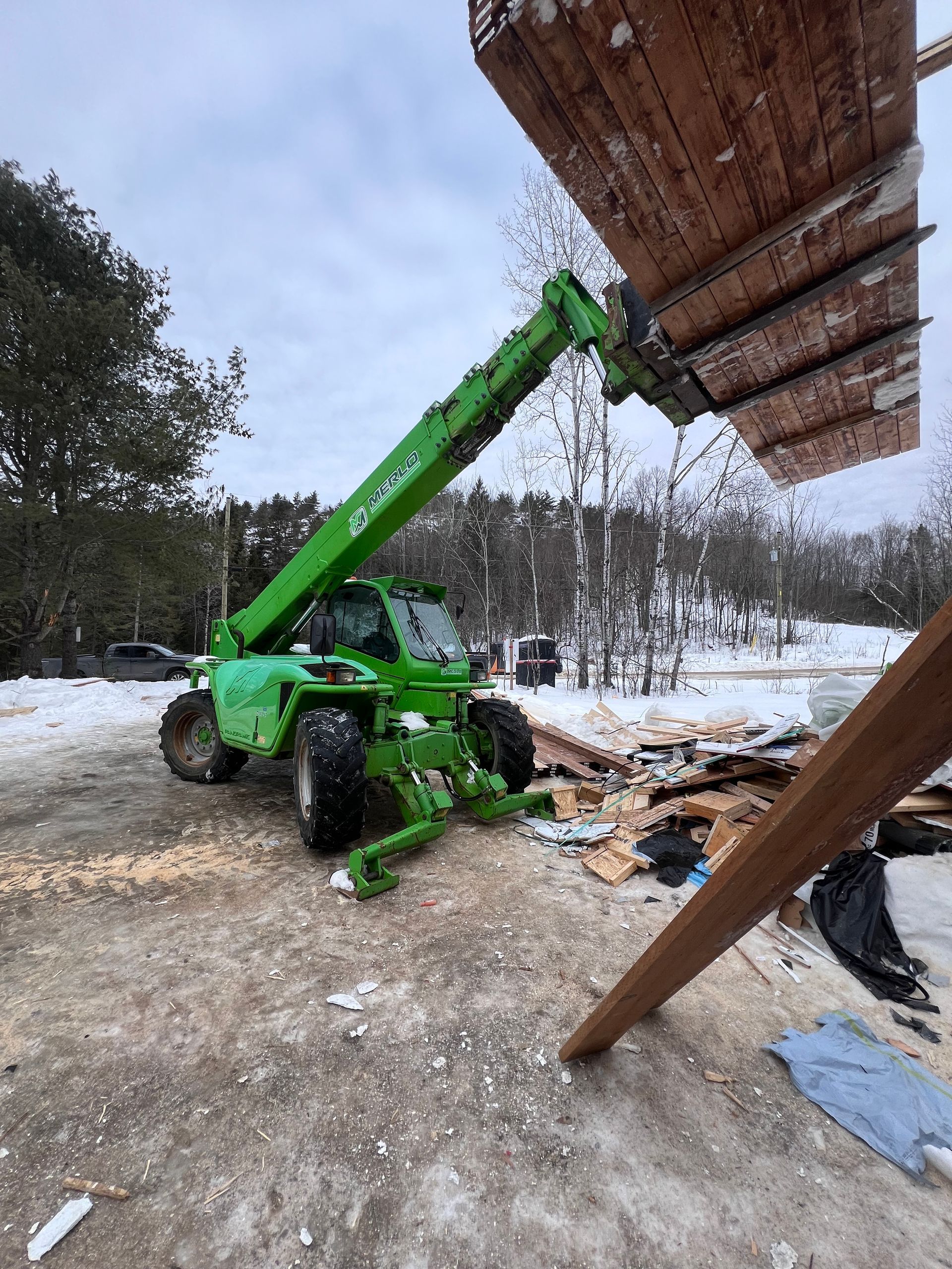 Un chariot télescopique vert soulève des planches de bois sur un chantier enneigé, à proximité de débris de construction.