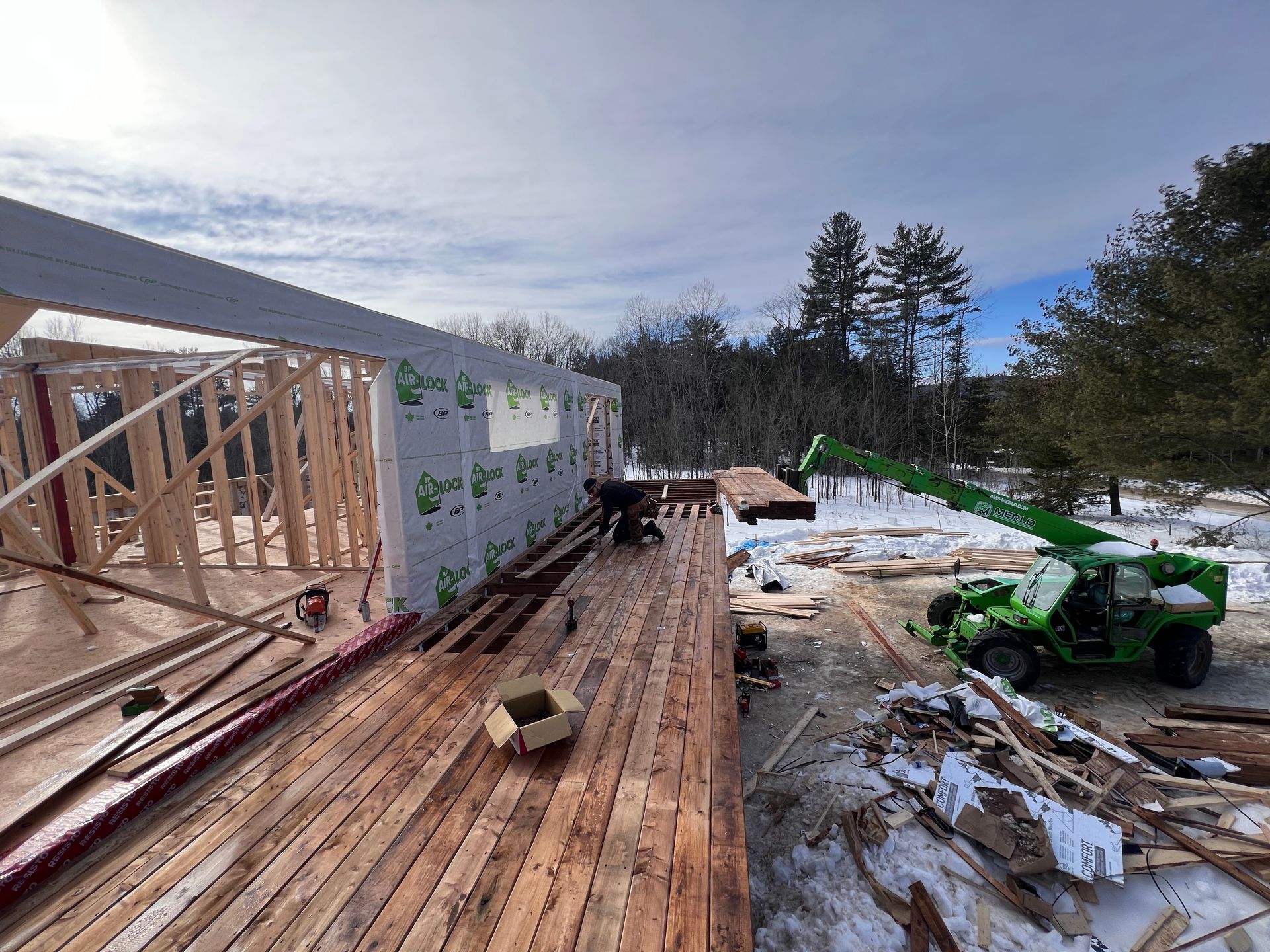Maison à ossature partiellement construite avec une terrasse en bois, neige au sol et un quad vert à proximité.