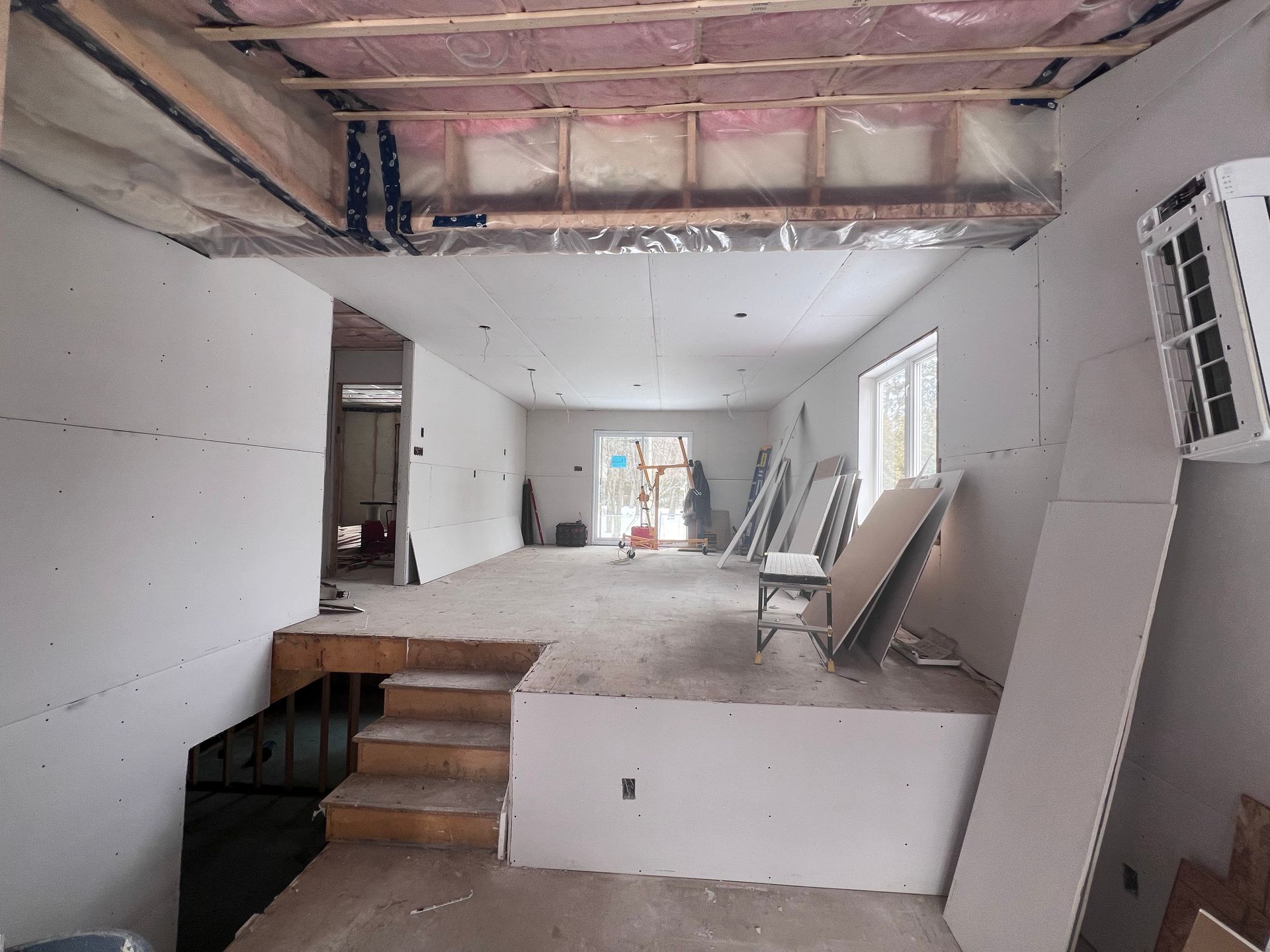 Pièce intérieure inachevée avec plafond apparent, cloisons sèches, marches et matériaux de construction près des fenêtres.