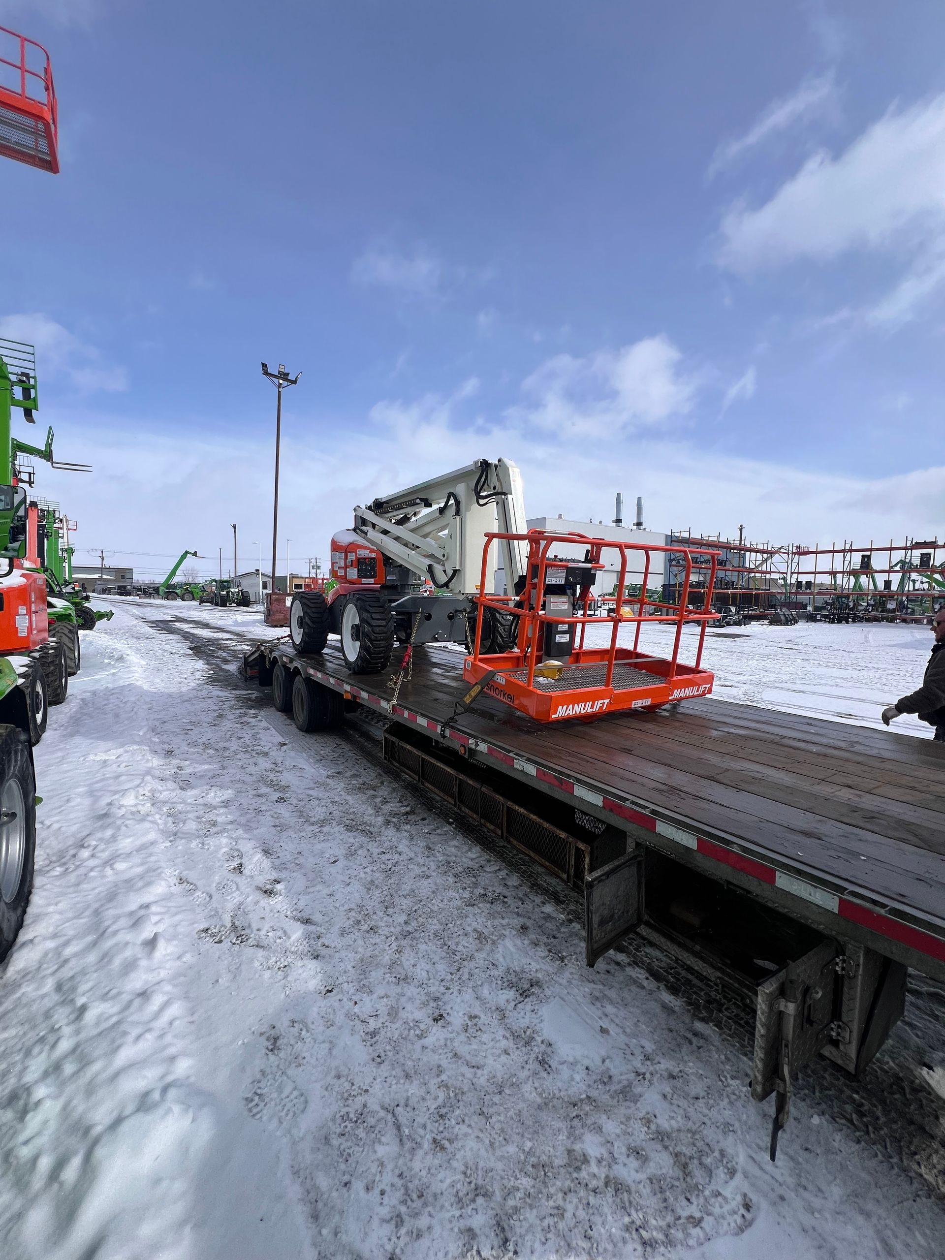 Cour industrielle enneigée avec une plateforme de travail orange et des machines sur une remorque à plateau sous un ciel bleu
