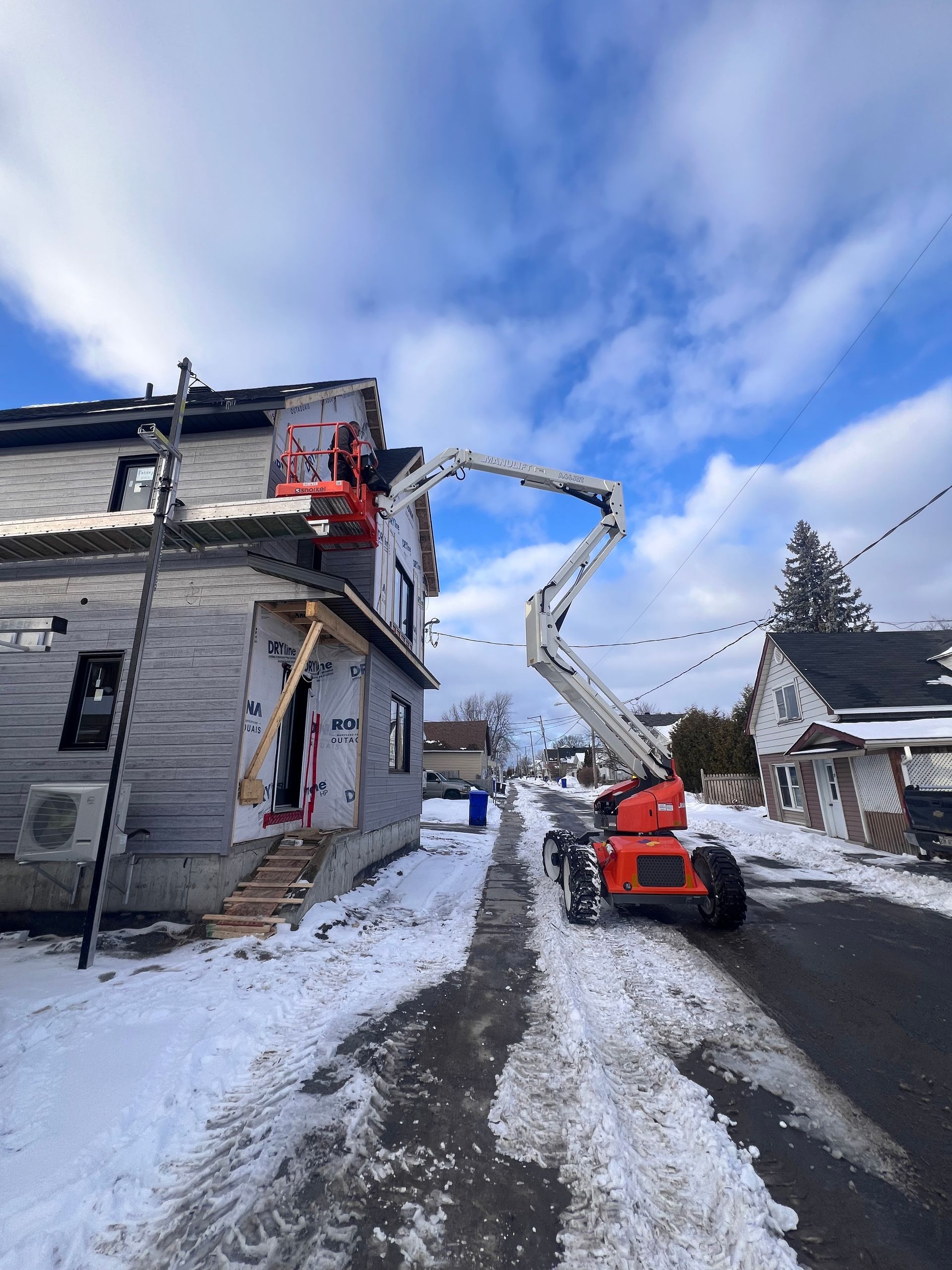 Une nacelle élévatrice orange dégage la neige près d'une maison grise dans une rue enneigée, sous un ciel bleu nuageux.