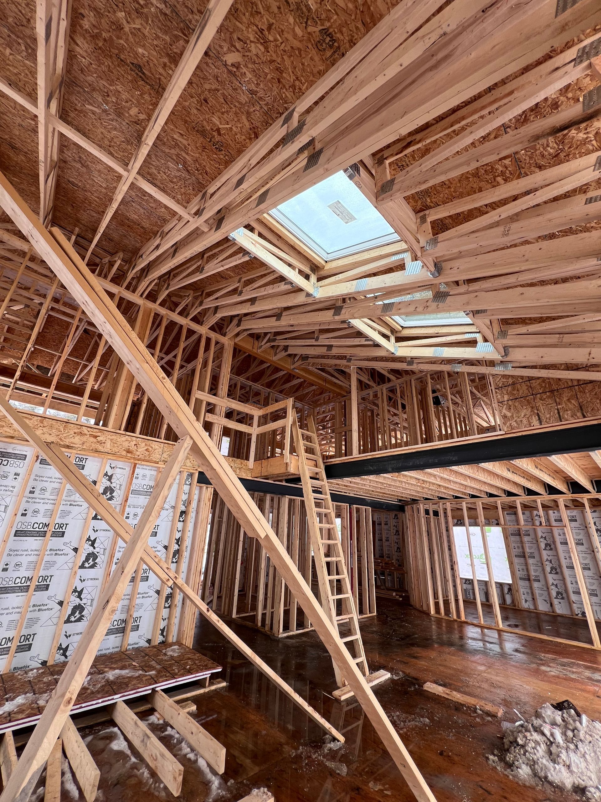 Intérieur d'une maison en bois en construction, avec charpente apparente, échelles et puits de lumière au plafond.