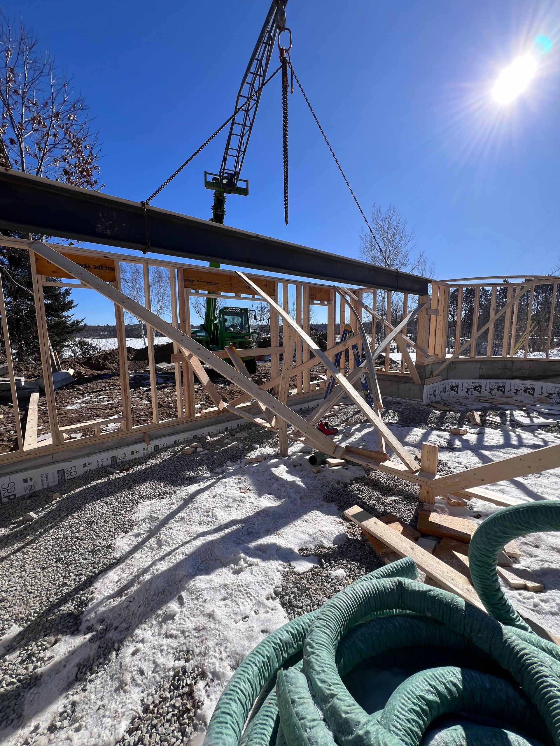 Chantier de construction avec charpente en bois, grue et tuyau d'arrosage vert enroulé sous un ciel d'un bleu éclatant