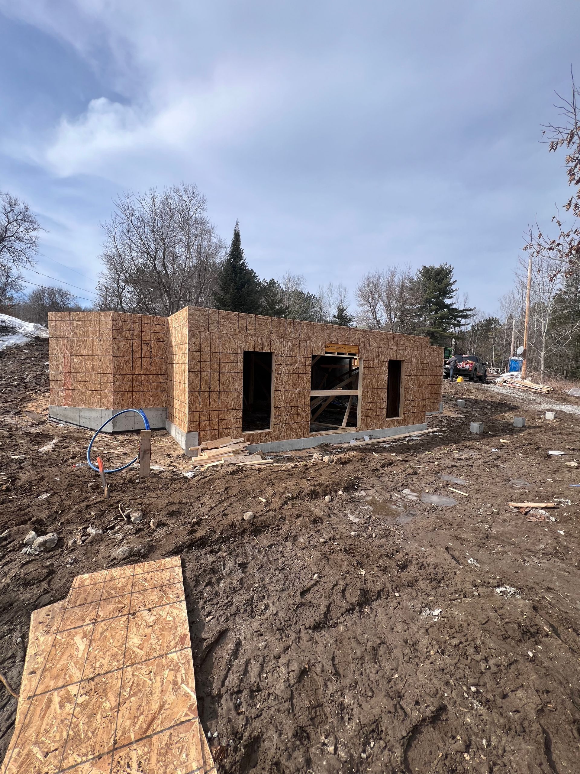 Structure en briques partiellement construite sur un chantier boueux, avec des panneaux de contreplaqué et des arbres dénudés à proximité.