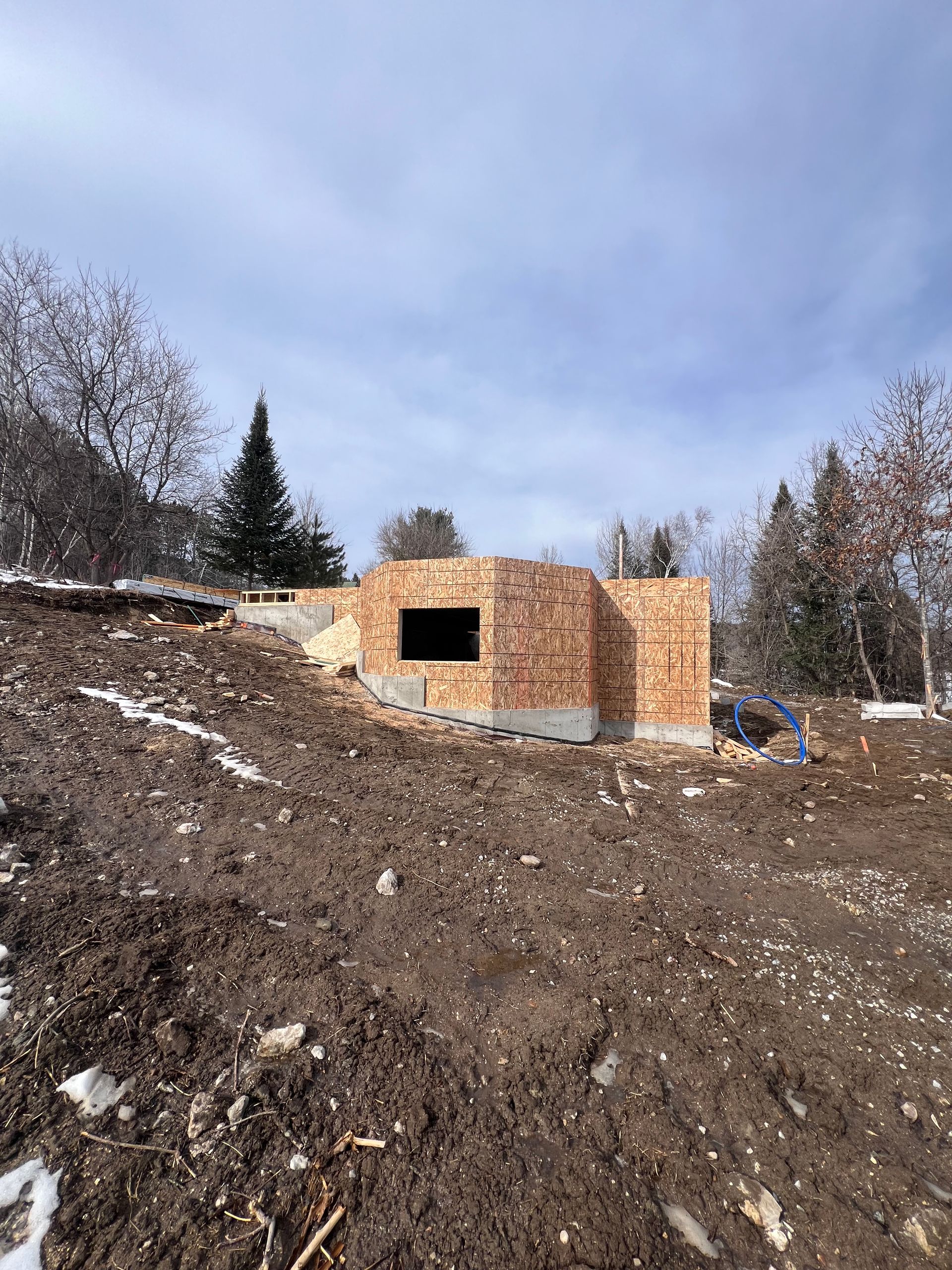 Petit bâtiment en bois inachevé sur un versant de terre enneigé sous un ciel nuageux, avec des arbres en arrière-plan