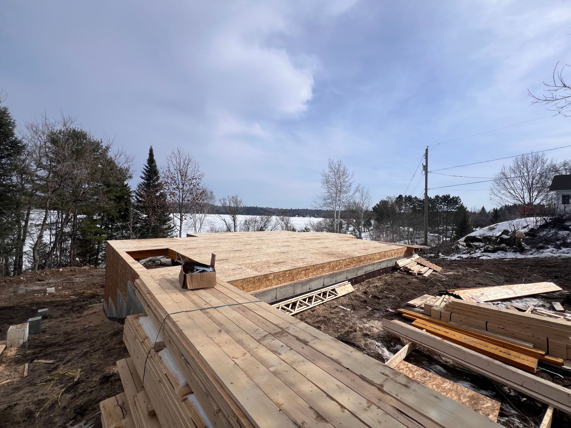 Fondations d'une maison en bois en construction sur un terrain dégagé, avec une charpente en bois et un ciel nuageux.