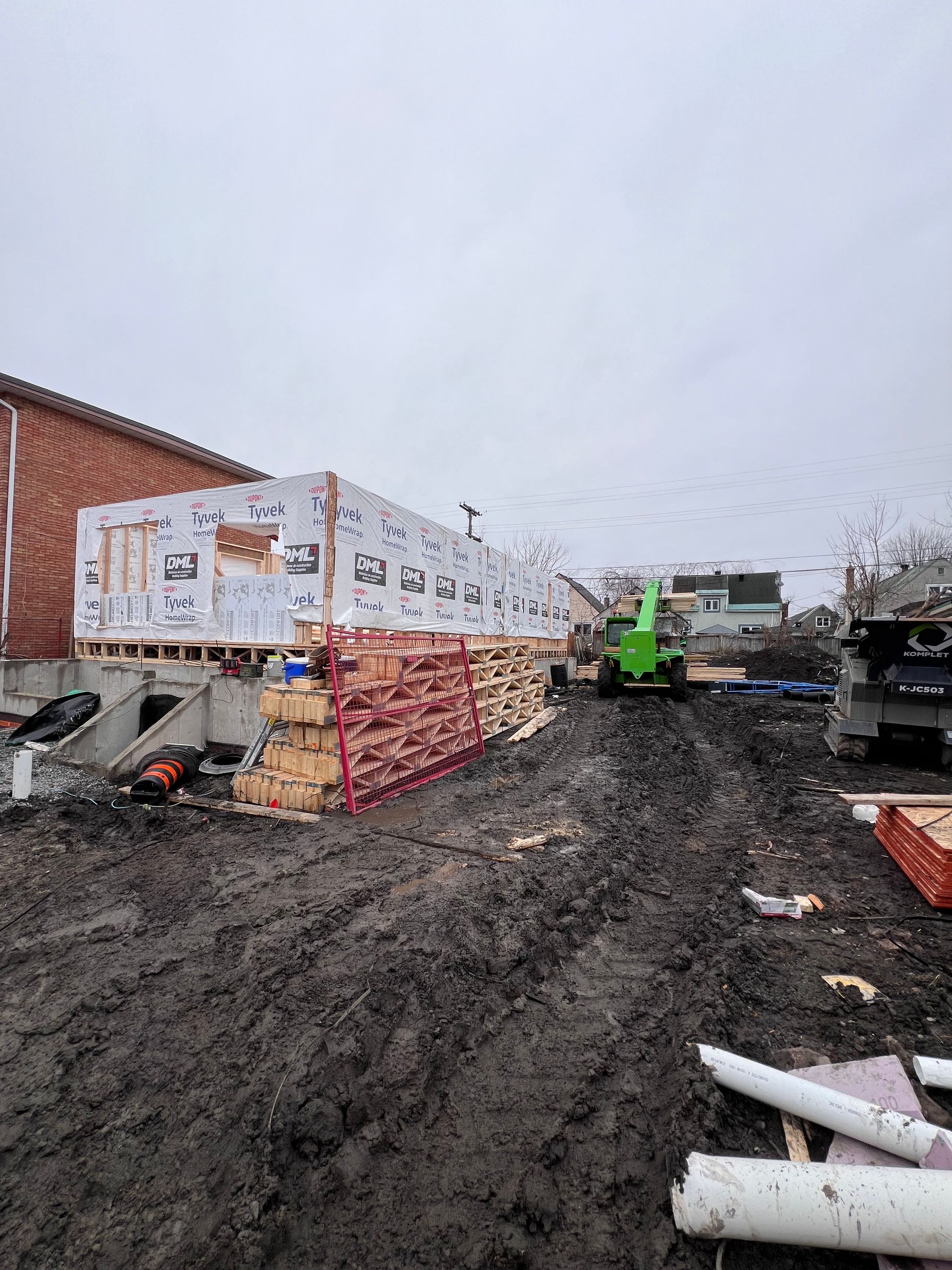 Chantier de construction avec sol boueux, matériaux empilés et un chariot élévateur vert à côté d'un bâtiment en briques.