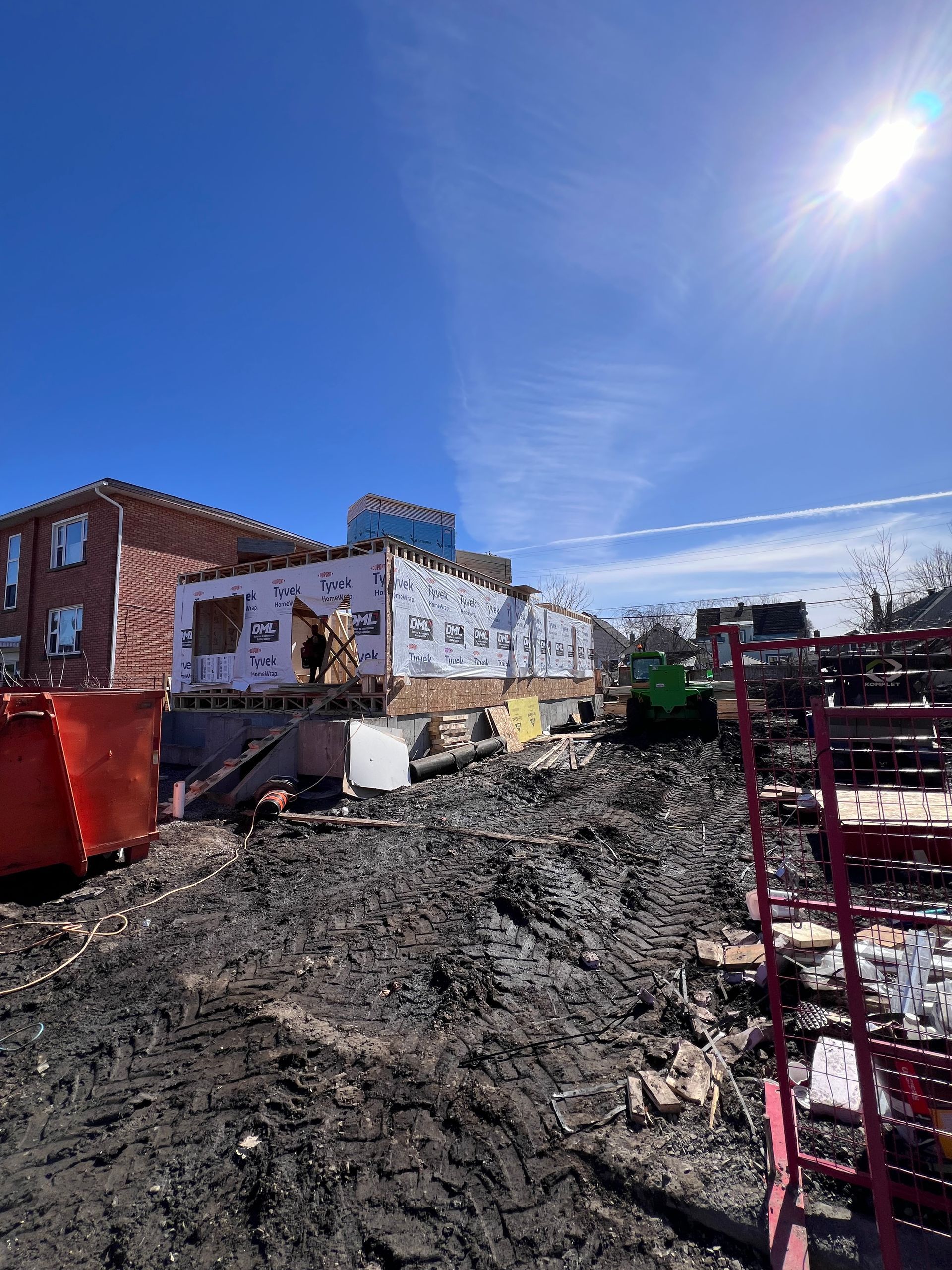Chantier de construction avec terrain boueux, bâtiment en briques blanches, engins de chantier et ciel d'un bleu éclatant