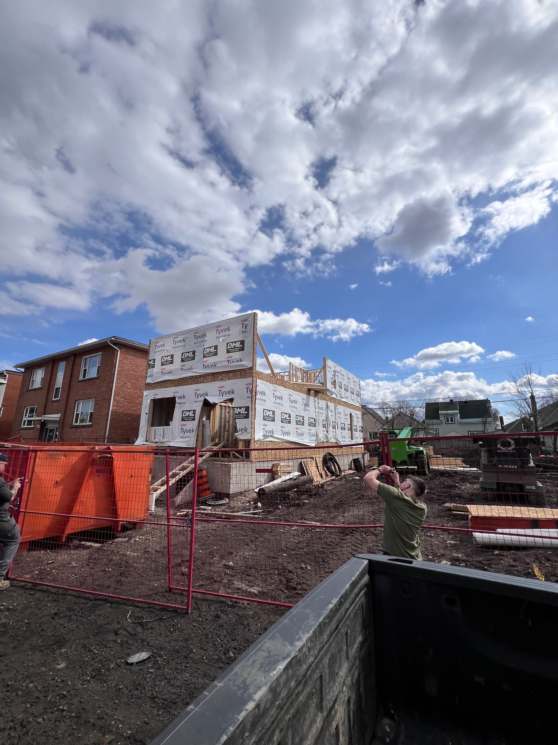 Chantier avec un bâtiment en briques partiellement démoli, une clôture orange et un ciel bleu nuageux.