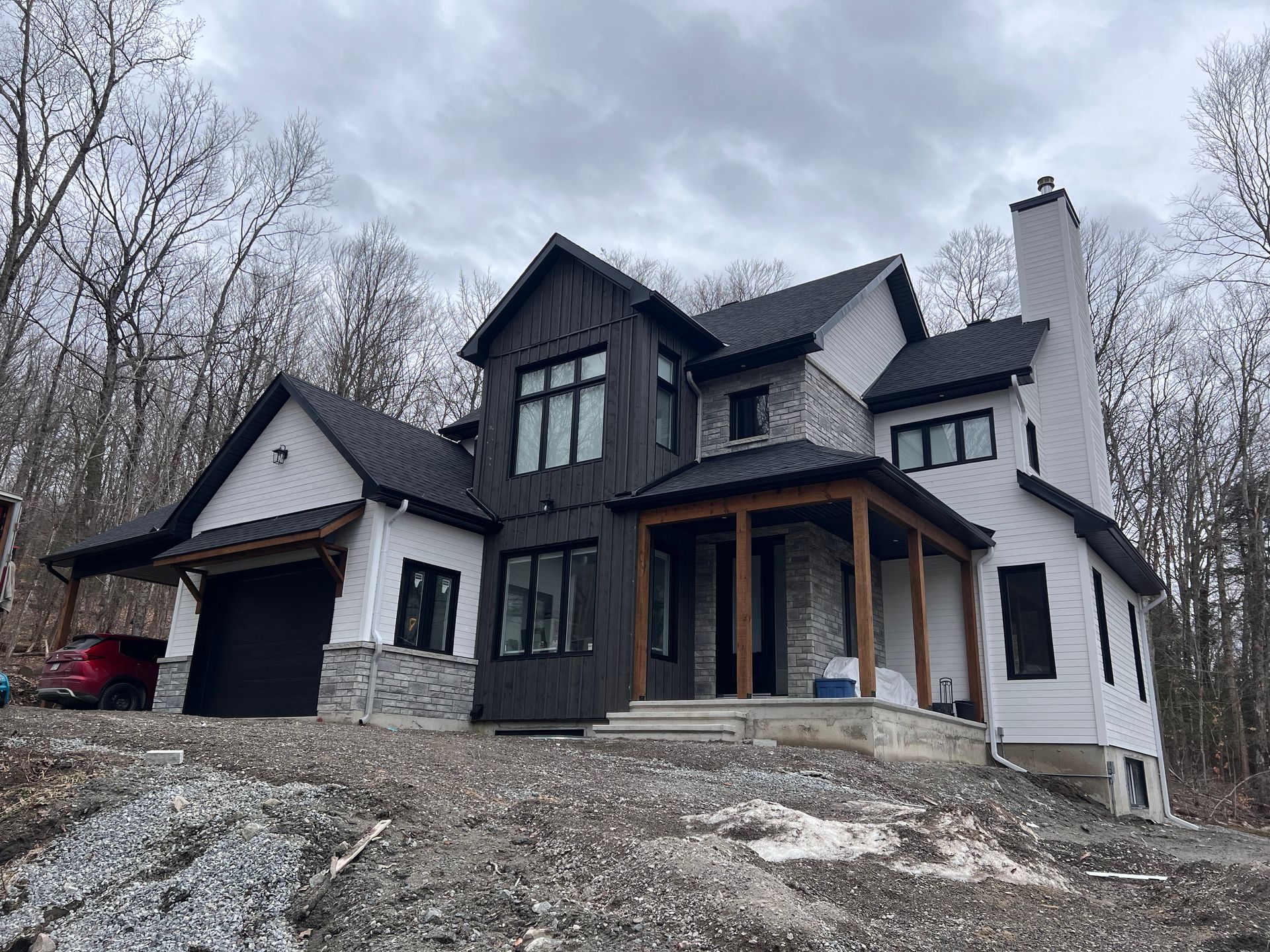 Maison moderne de deux étages en construction, avec des boiseries sombres, un bardage blanc et des arbres dénudés sur un terrain en pente.