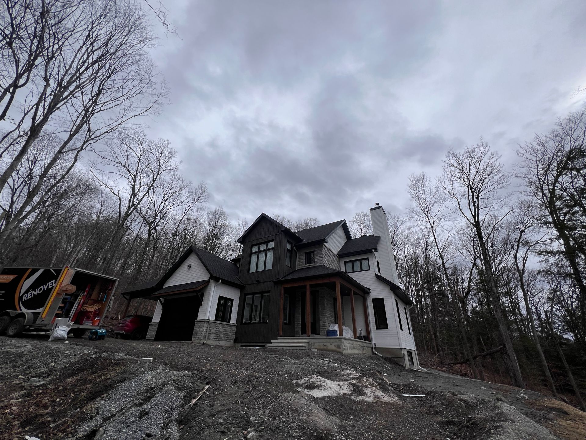 Grande maison blanche au toit sombre sur un flanc de colline boisé sous un ciel nuageux