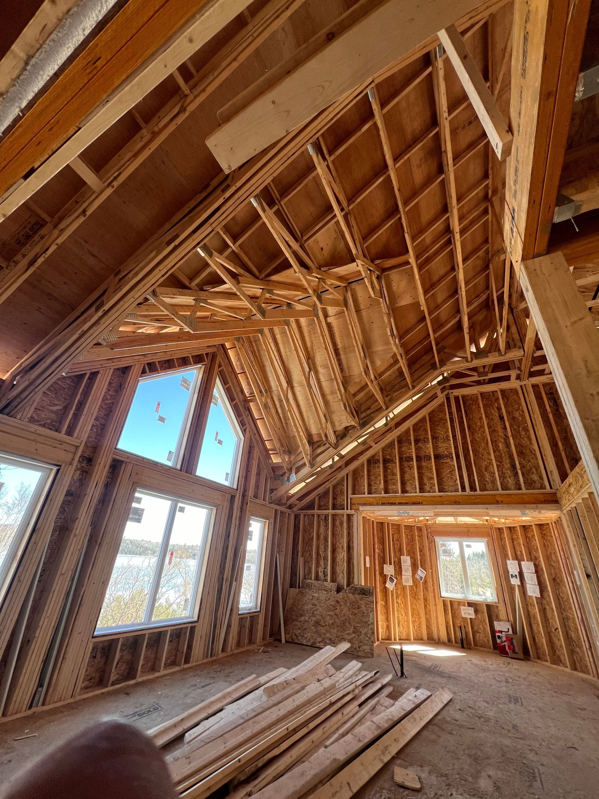 Intérieur d'une maison à ossature bois inachevée, avec charpente apparente et piles de bois au sol.