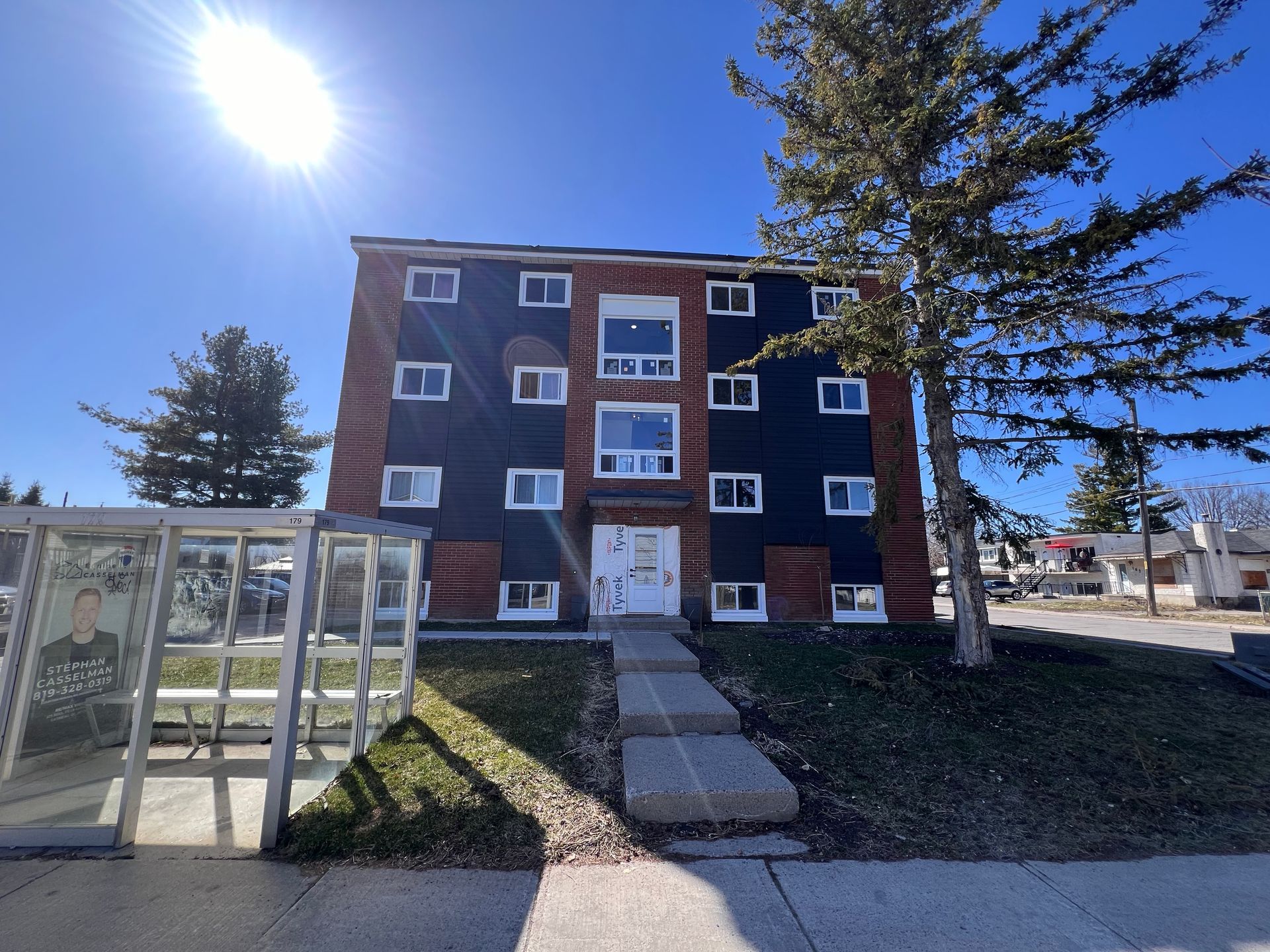 Immeuble d'appartements en briques avec entrée centrale, allée et abribus vitré sous un soleil éclatant