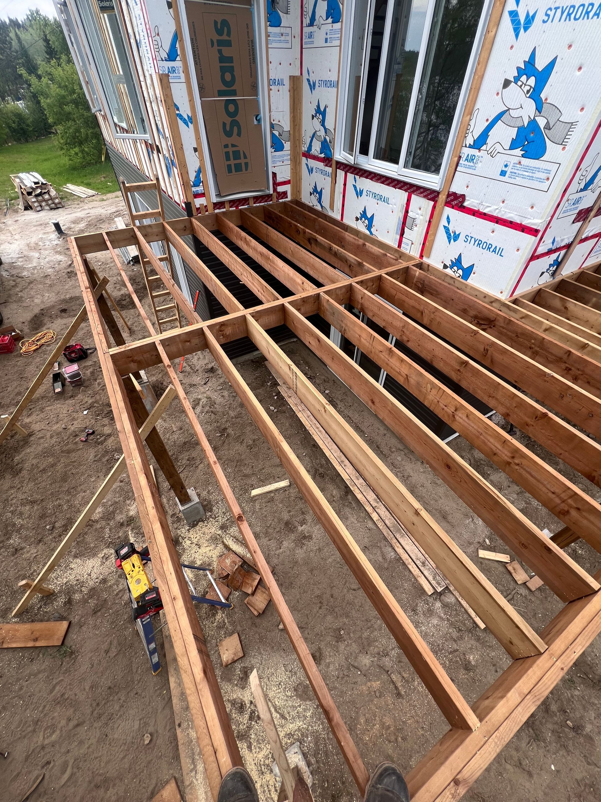 Solives de plancher en bois en cours de construction à côté d'un mur de maison à ossature bois et de fondations en terre.