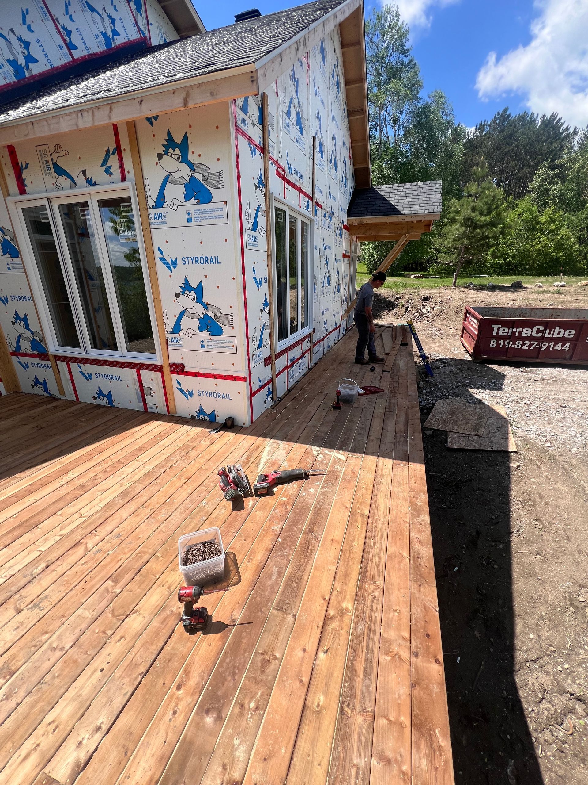 Maison en construction à bardage bois, peinture écaillée, terrasse en bois, outils et tranchée creusée à côté.