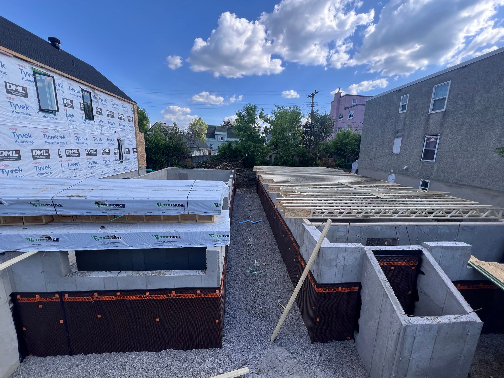 Chantier de construction avec charpente de maison, bardage blanc et terrasse sur le toit entre deux bâtiments sous un ciel bleu