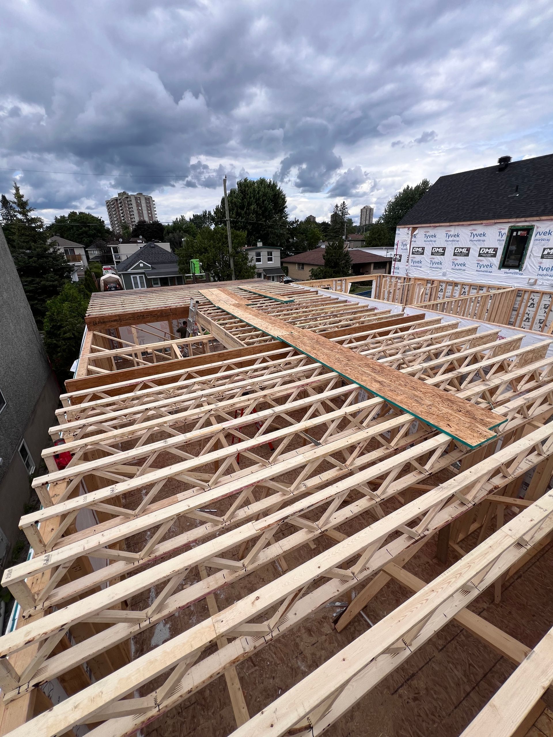 Charpente de toit en construction sur une maison, avec fermes en bois apparentes sous un ciel nuageux.