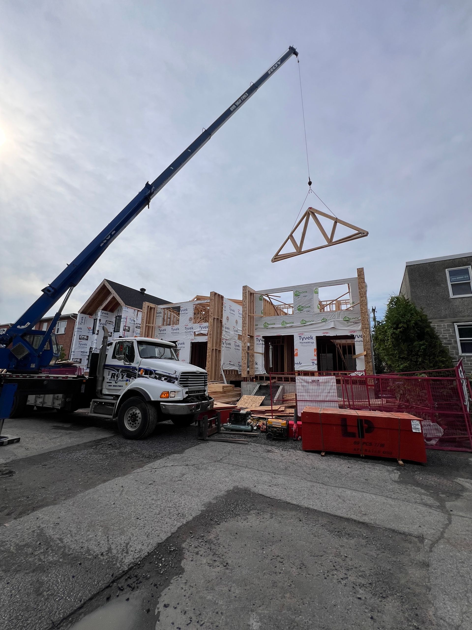 Une grue soulève une ferme de toit en bois sur un chantier de construction, avec un camion et du matériel de chantier.