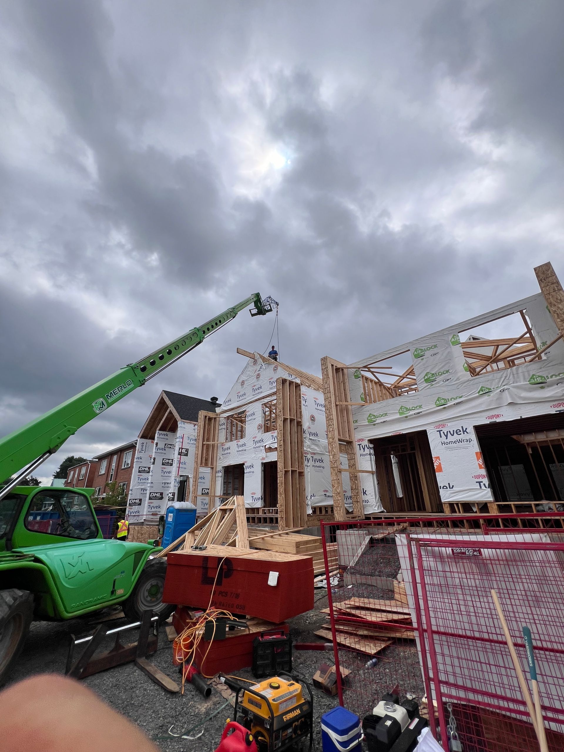Chantier de construction avec nacelle élévatrice verte à côté d'une maison en briques partiellement construite sous un ciel nuageux